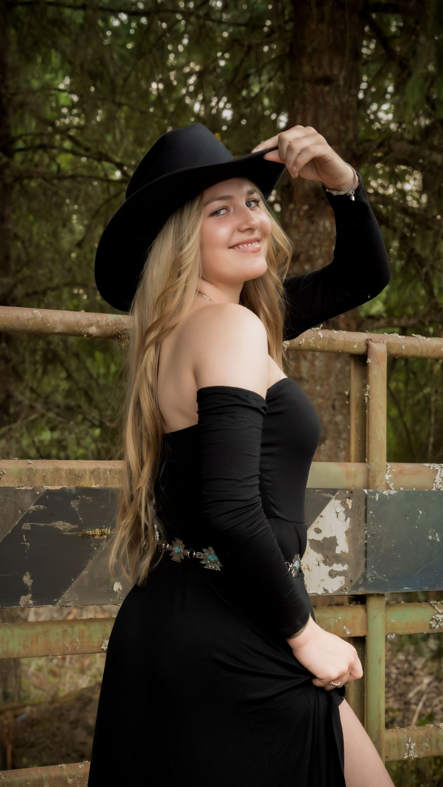 Young blonde woman in black strapless gown and wide-brim hat poses outdoors by rustic fence.