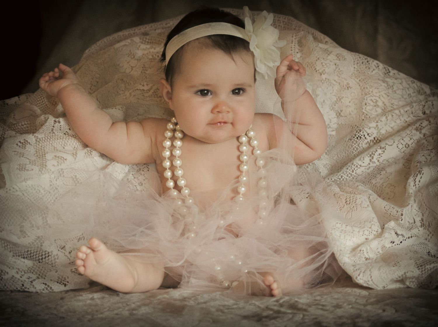 Adorable baby girl in white tutu dress and pearl necklace with headband, posed elegantly on lace fabric.
