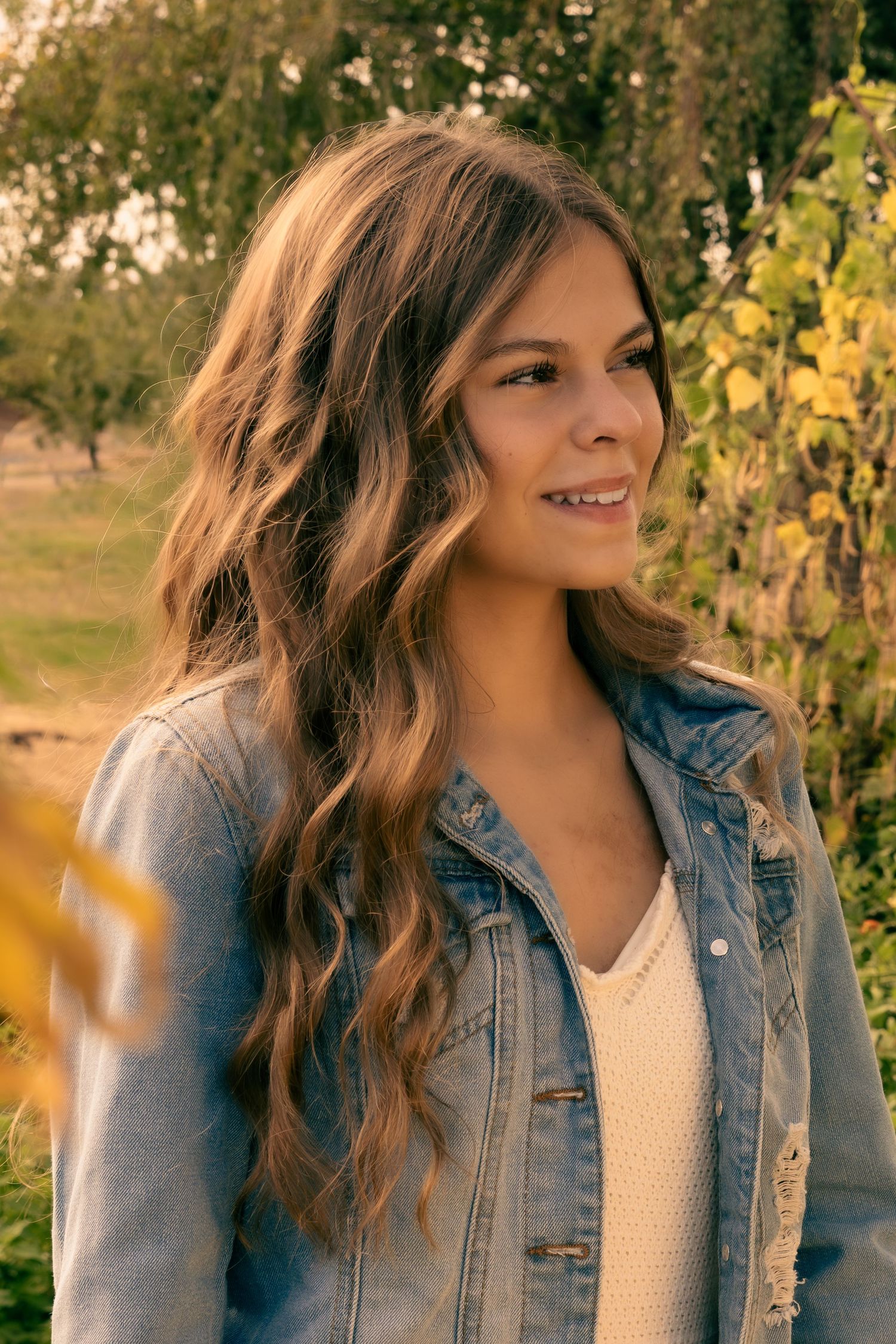 Young woman with long wavy hair in a denim jacket smiling outdoors among green and yellow autumn foliage.