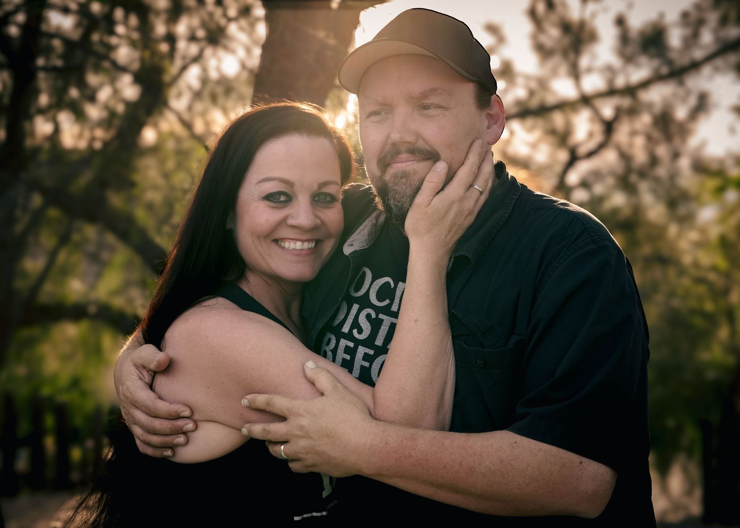 A smiling couple embraces outdoors at golden hour, woman in black top hugging bearded man in dark t-shirt. and hat. moody authentic embrace