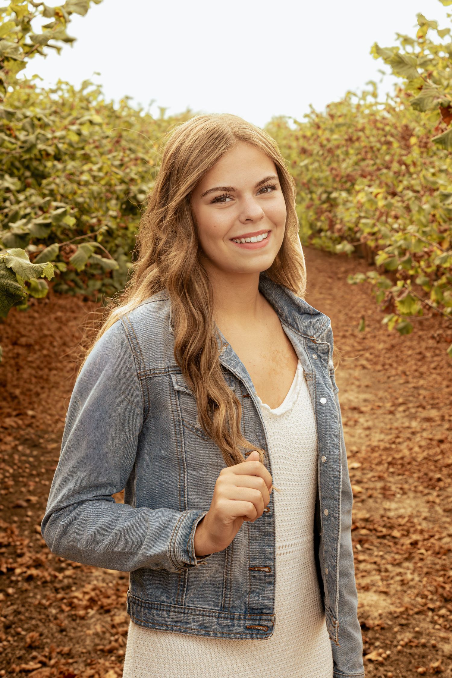 Young woman in denim jacket and white dress smiling in an orchard pathway surrounded by green foliage.