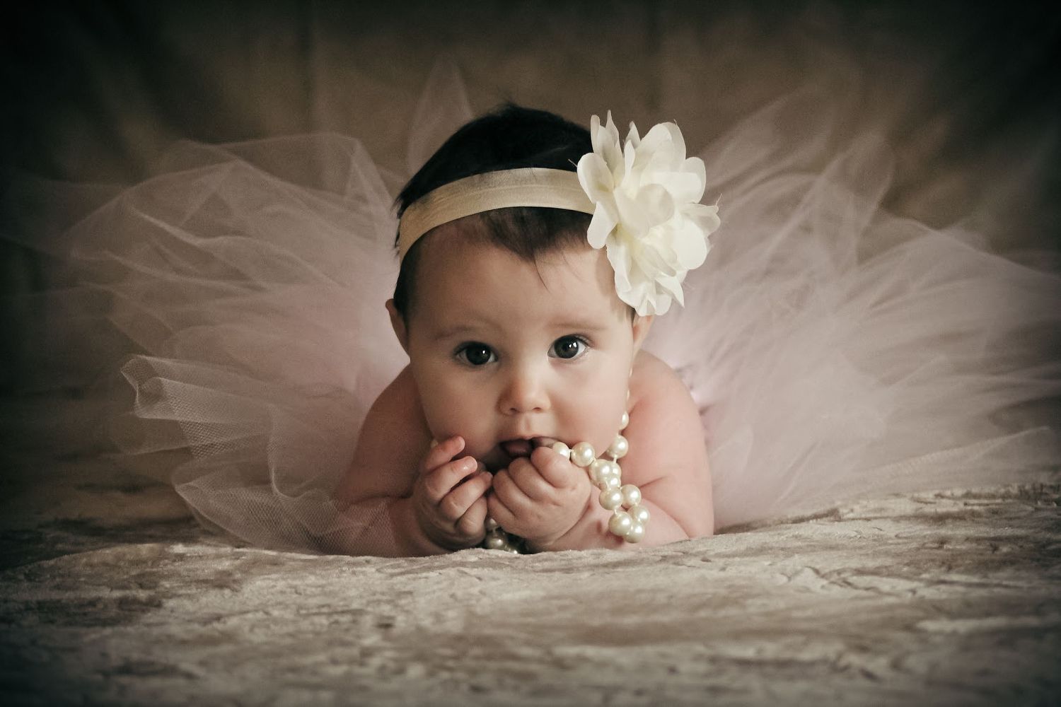 Adorable baby girl in pink tutu and floral headband chewing pearl necklace while posing on lace fabric.