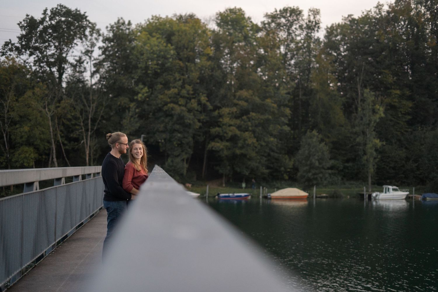 Wedding photo shoot on a lakeside dock with boats and trees in the background during an overcast day.