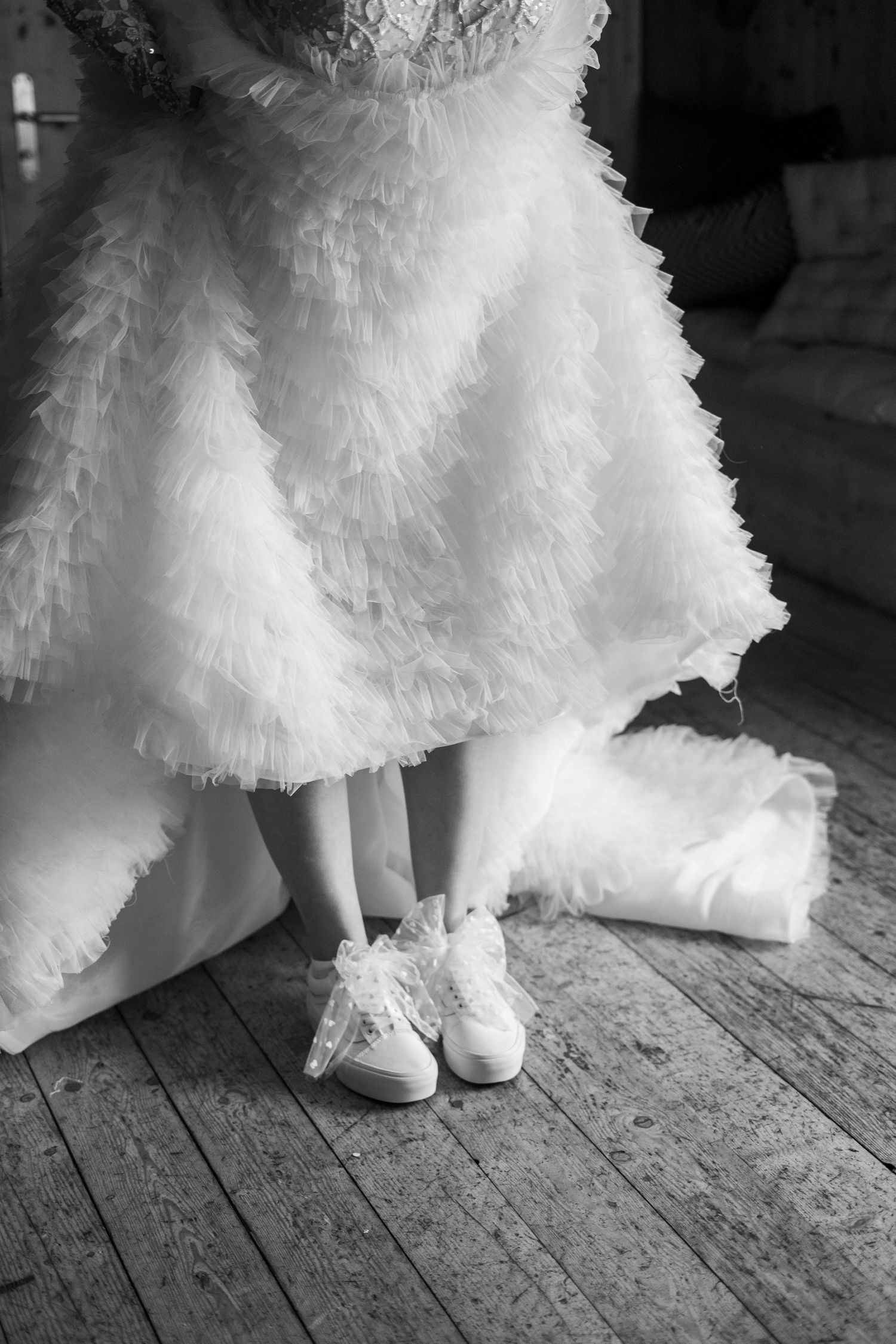 Close-up of white sneakers worn with a ruffled wedding dress on a wooden floor.