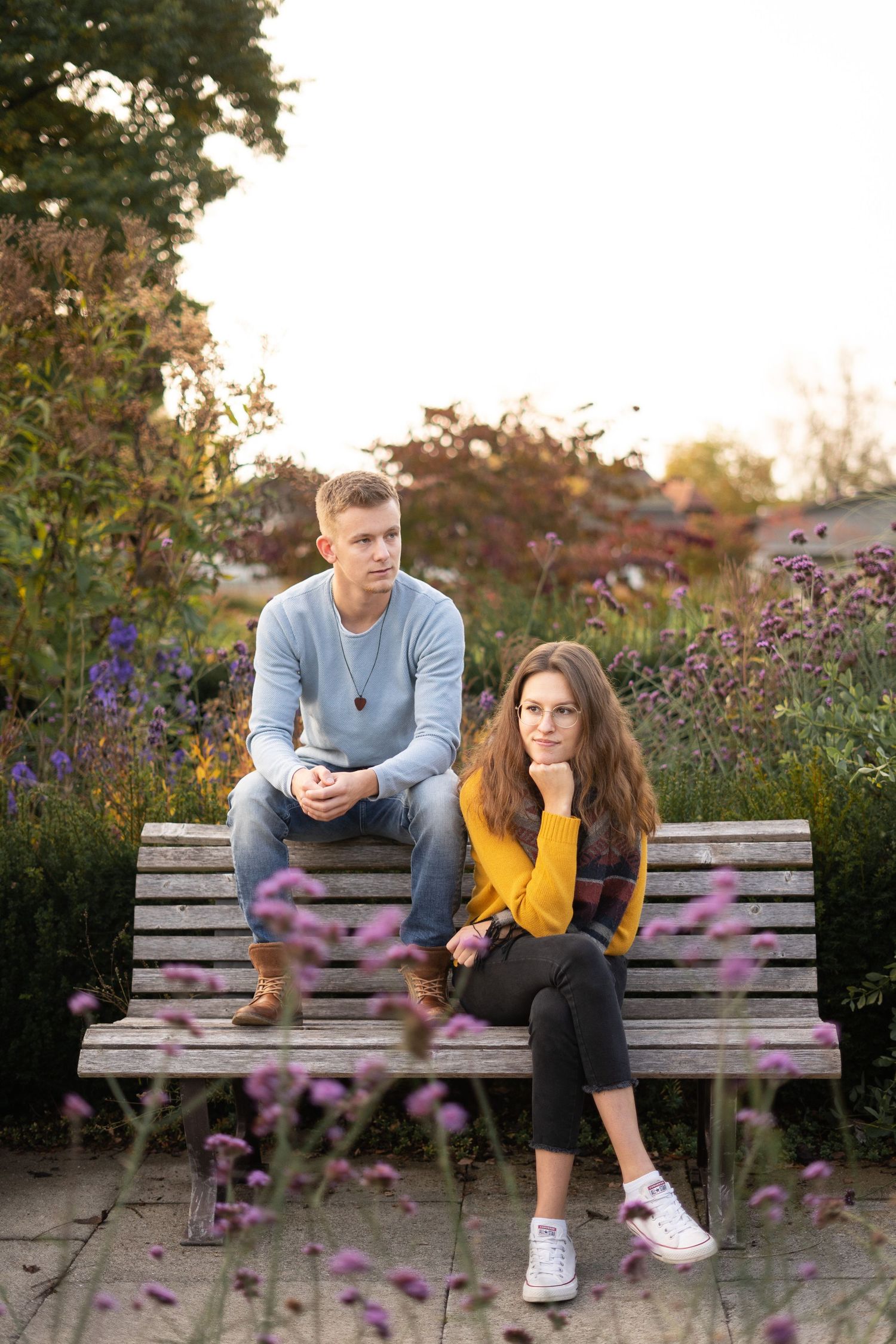 A couple sitting pensively on a wooden bench surrounded by purple flowers in a garden setting.