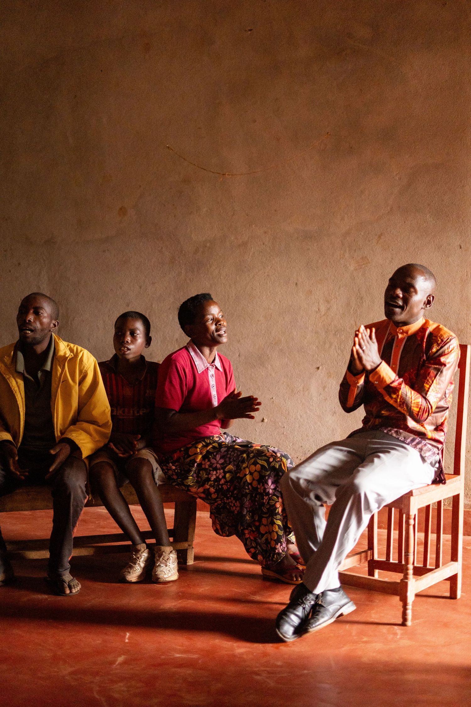 Four people sitting and clapping on wooden benches against an earthen wall.