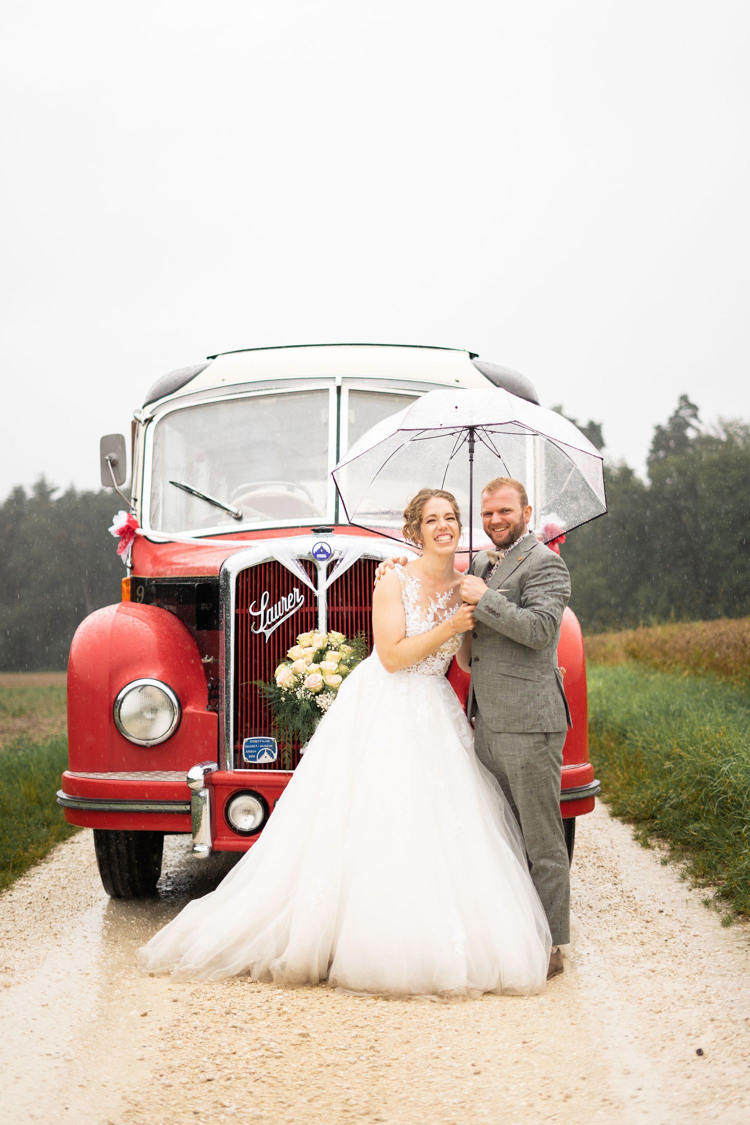 A newlywed couple poses with a vintage red bus on a country road while holding a white umbrella.