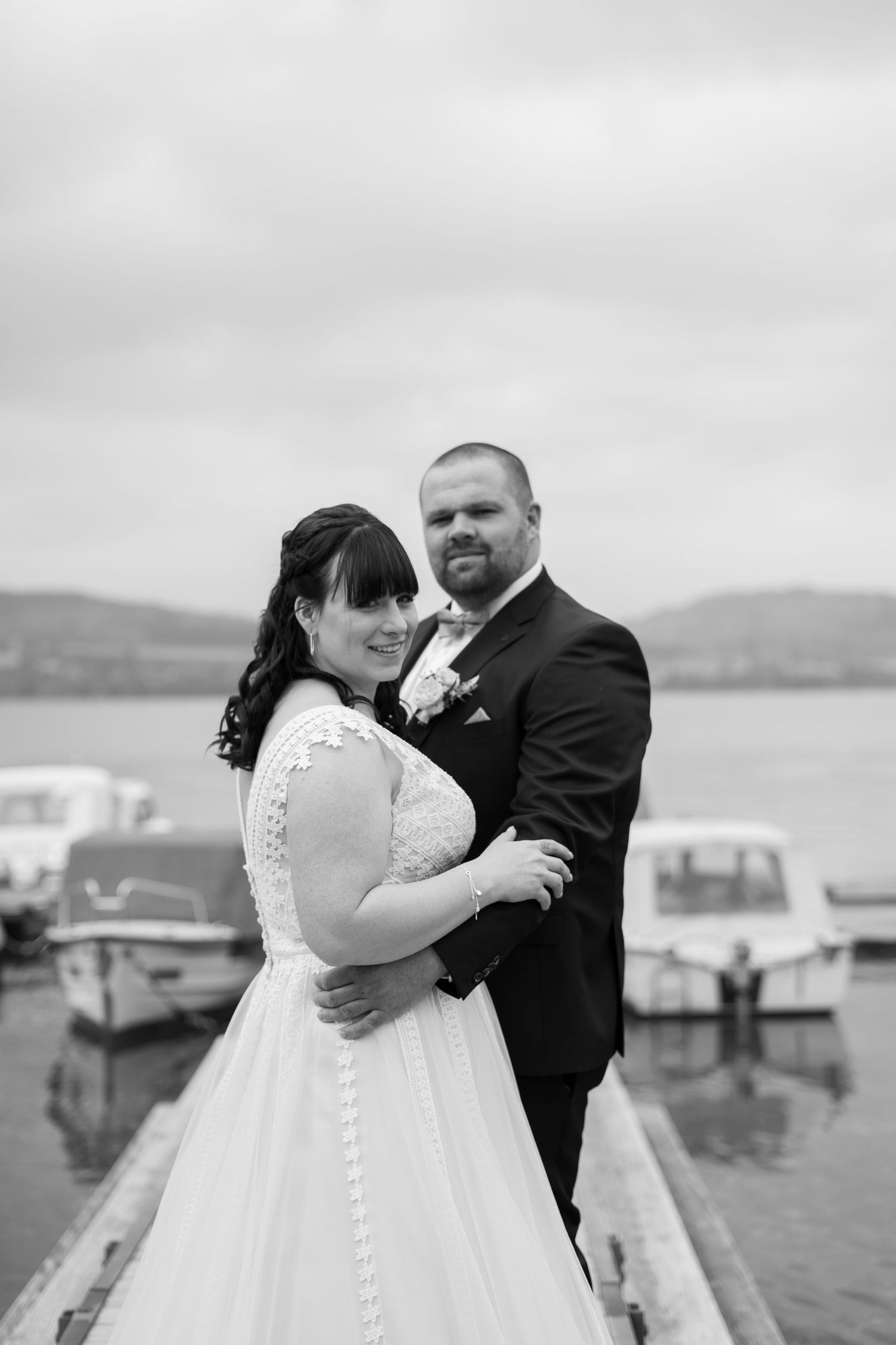 Wedding couple embracing on a wooden dock with boats and mountains visible in the background.