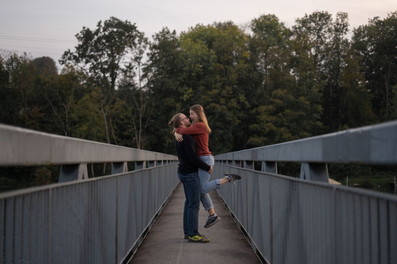 A couple embraces on a metal footbridge during sunset with trees in the background.