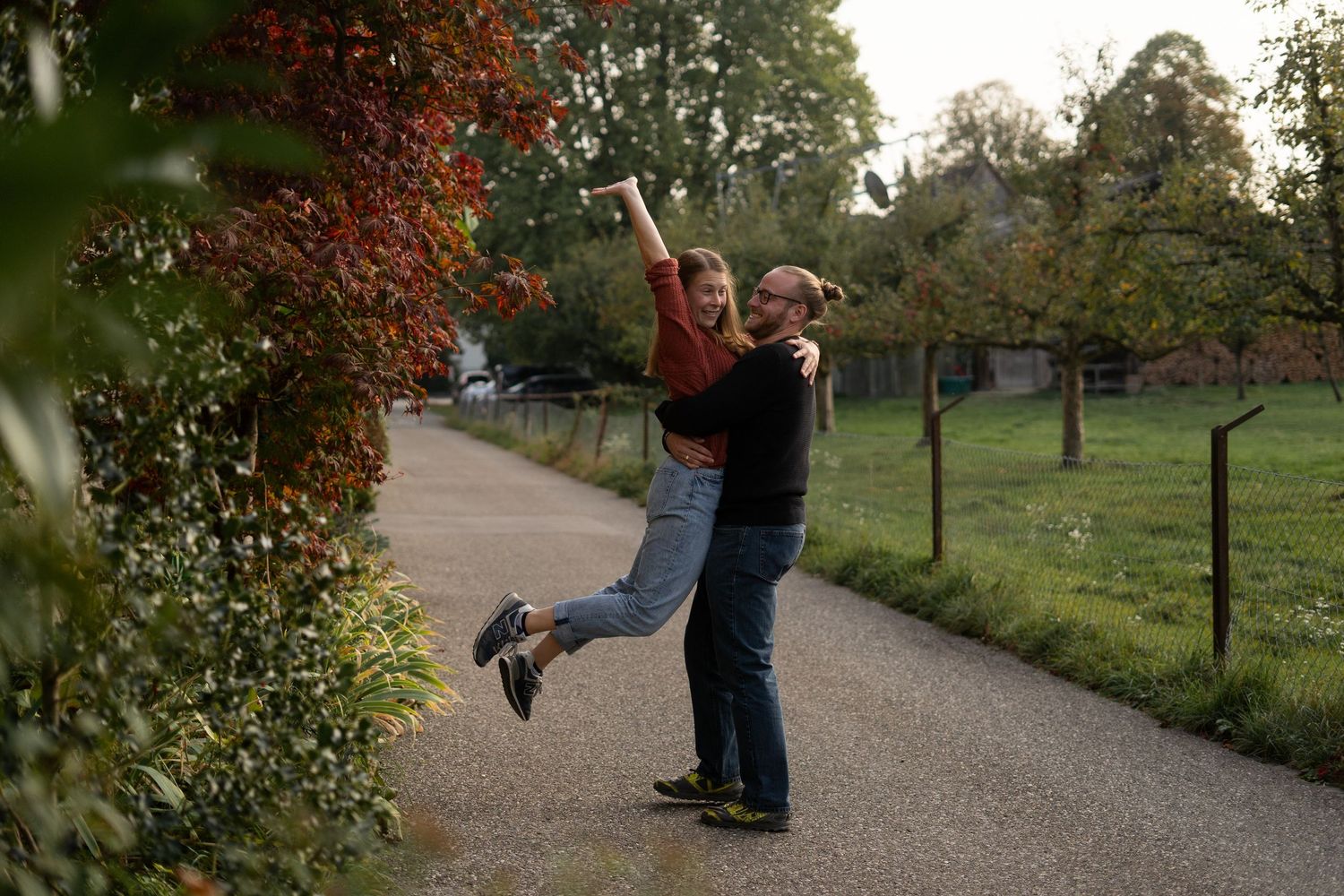A couple shares a playful lift moment on a paved path surrounded by autumn trees.