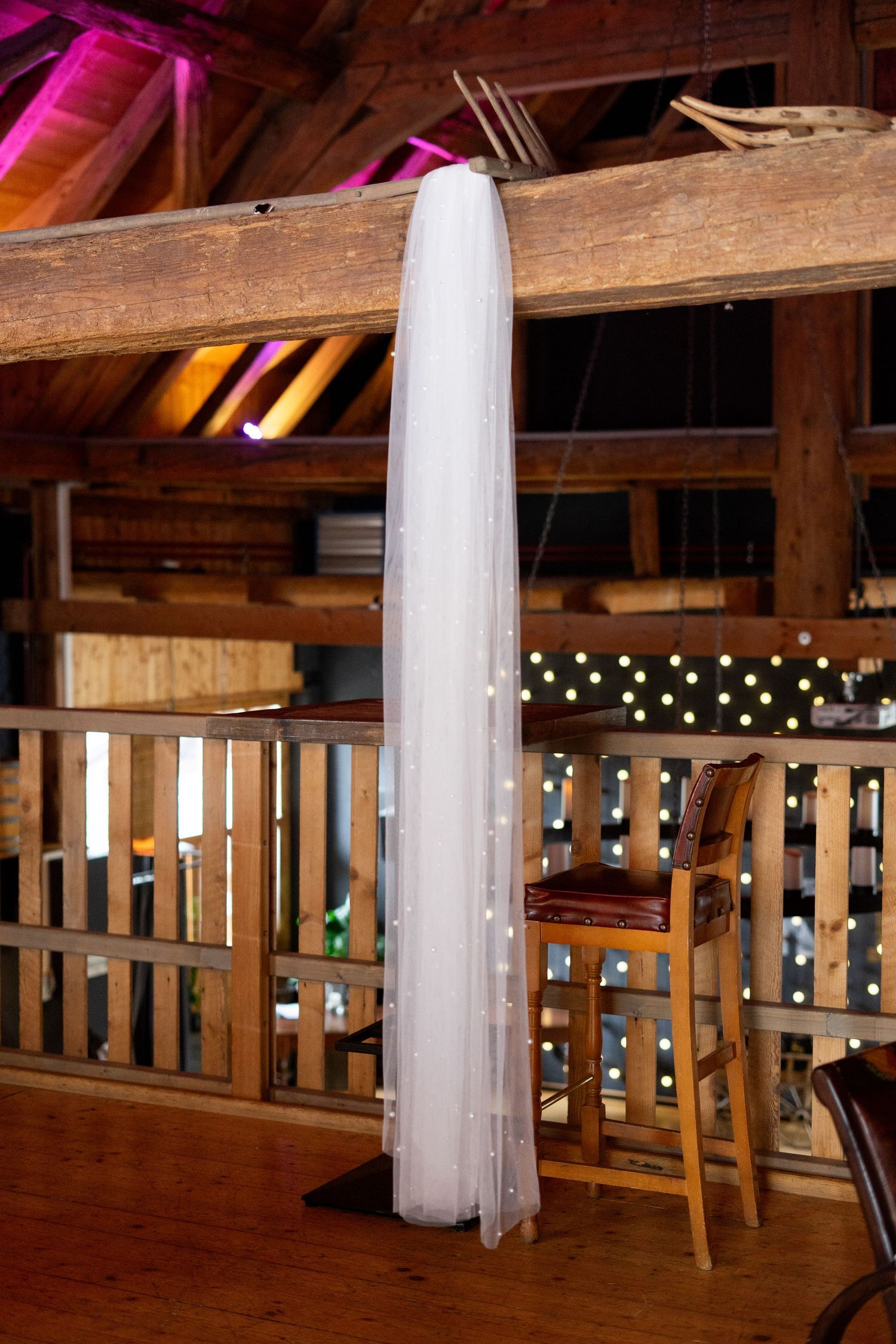 Long white wedding veil hanging from wooden beam in rustic barn venue with string lights.