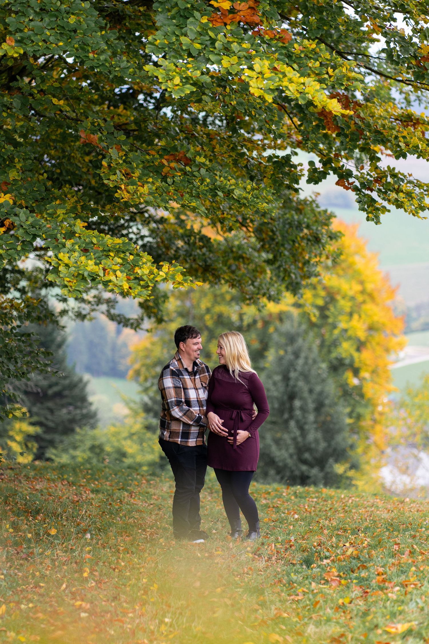 A couple shares a tender moment under vibrant autumn trees with golden leaves falling around them.