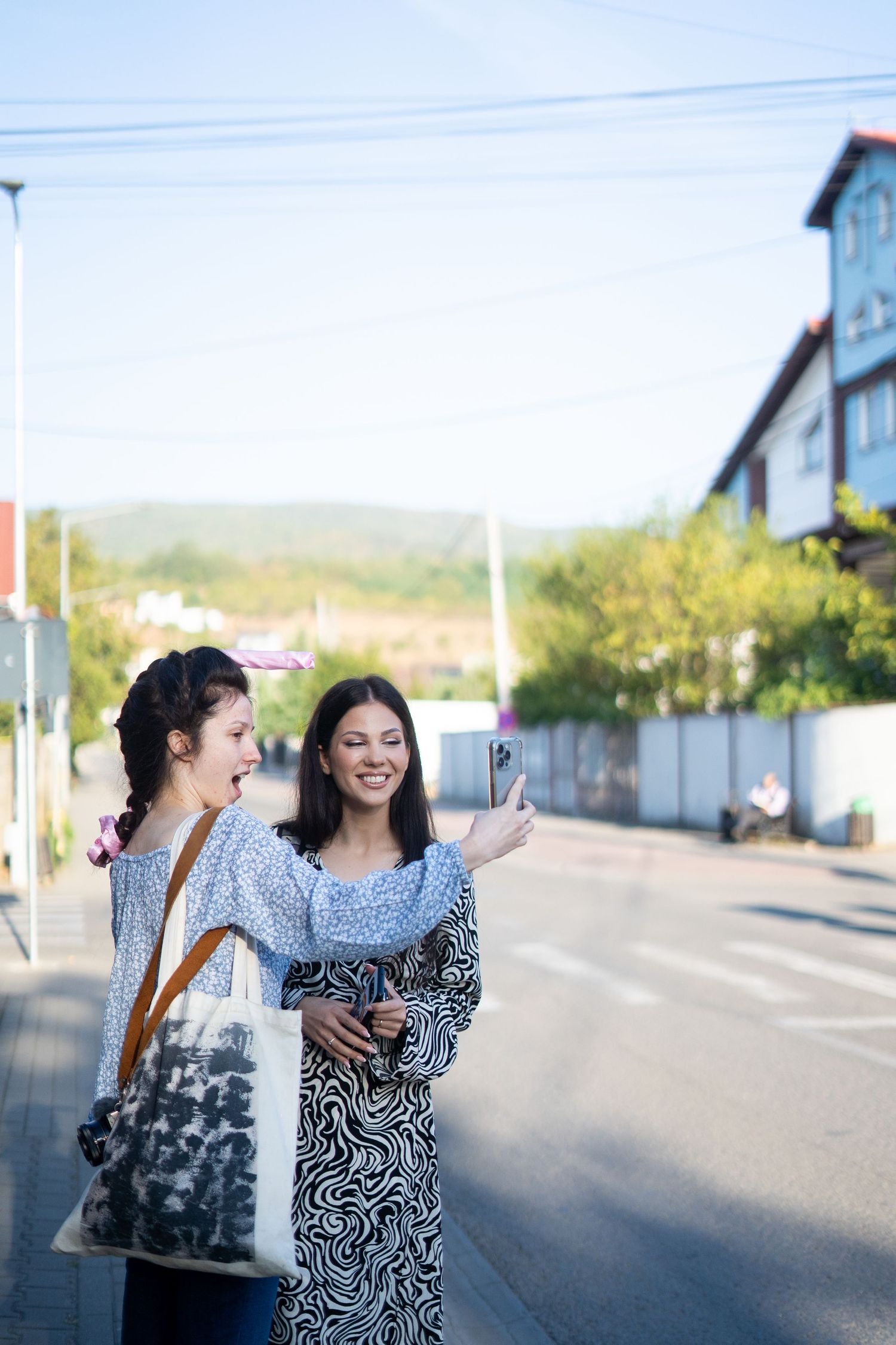 Two friends taking selfies together on a sunny street with buildings in the background.