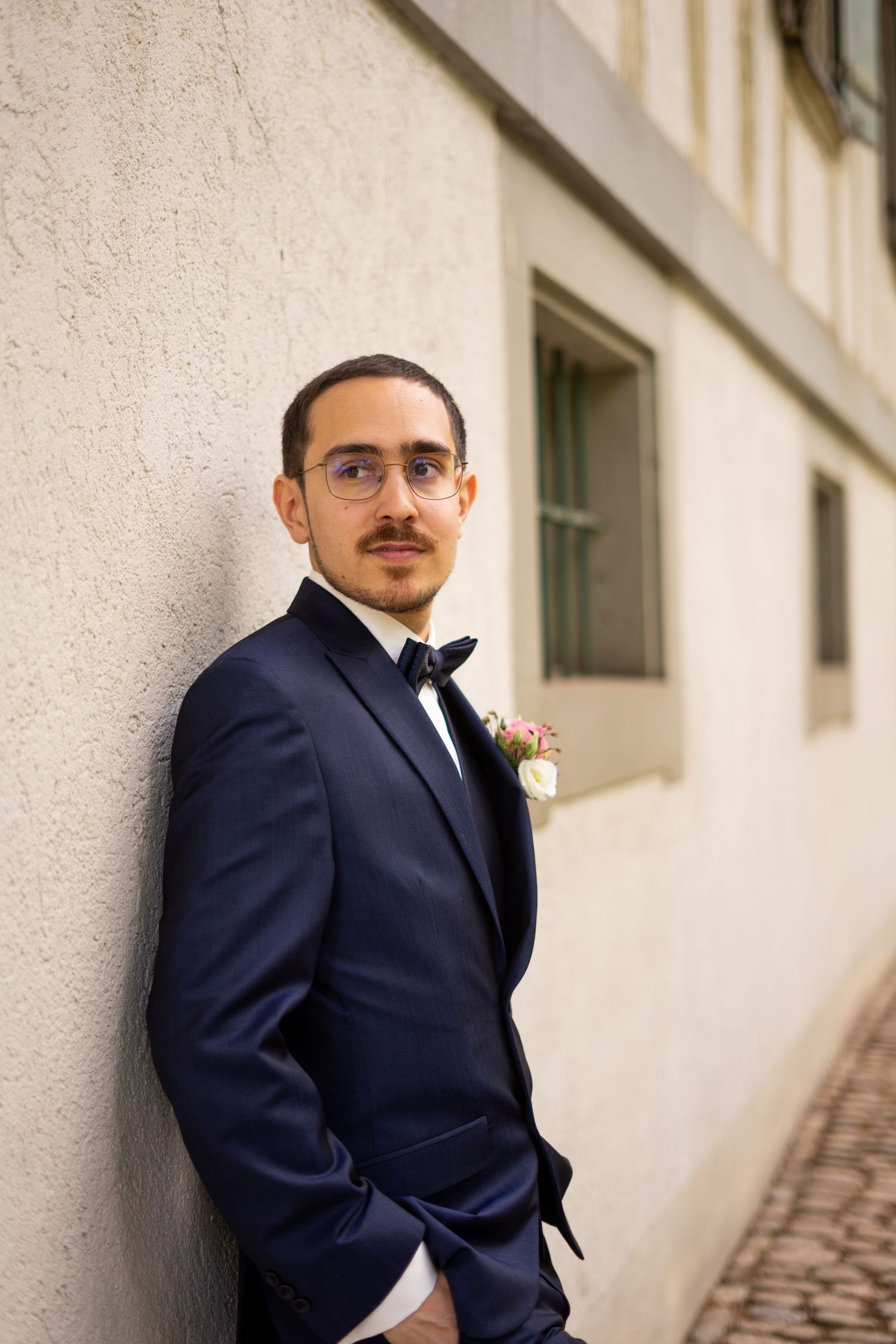 A groom in a navy tuxedo and bowtie poses against a light colored wall.