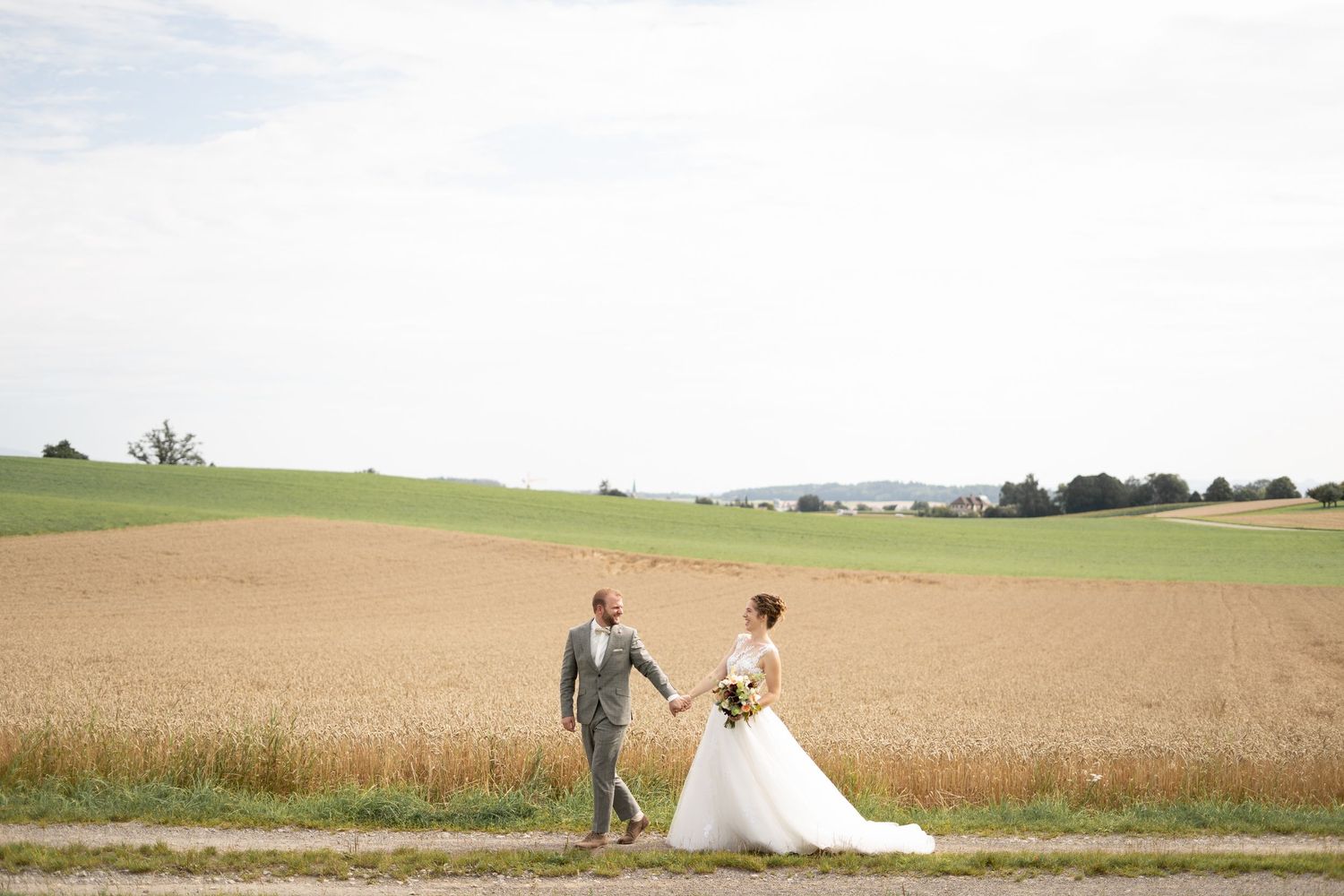 A bride and groom walk hand in hand through a golden wheat field under cloudy skies.