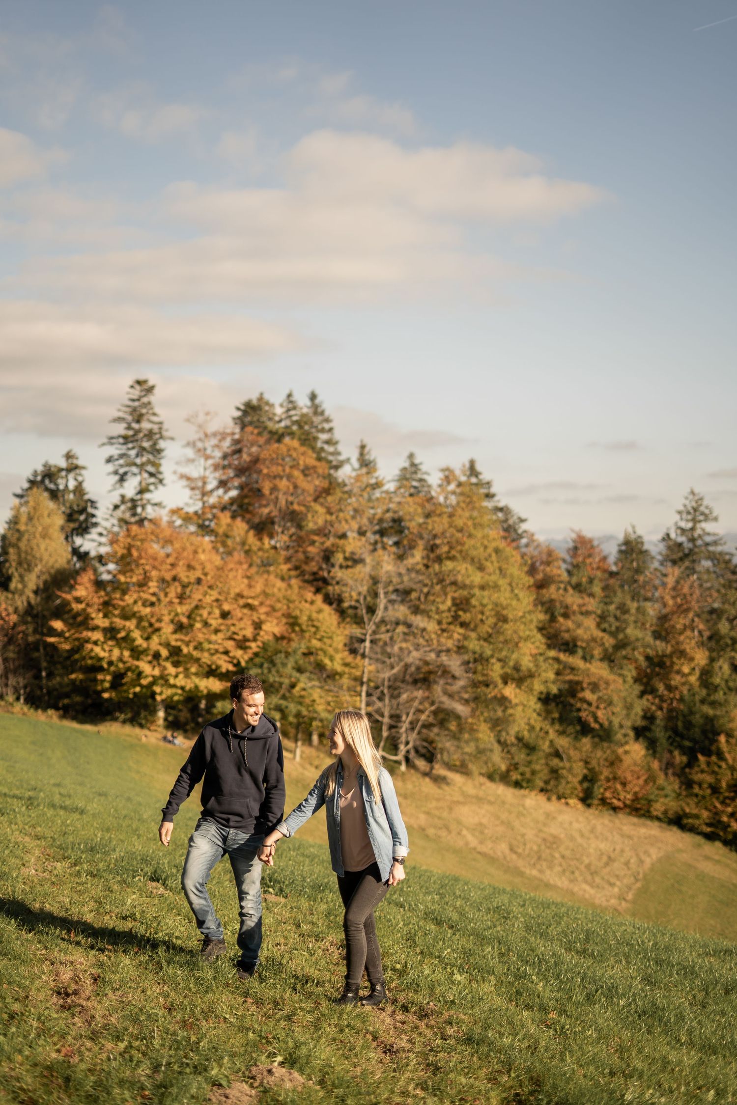 A couple walks hand in hand through a hillside meadow surrounded by autumn trees.