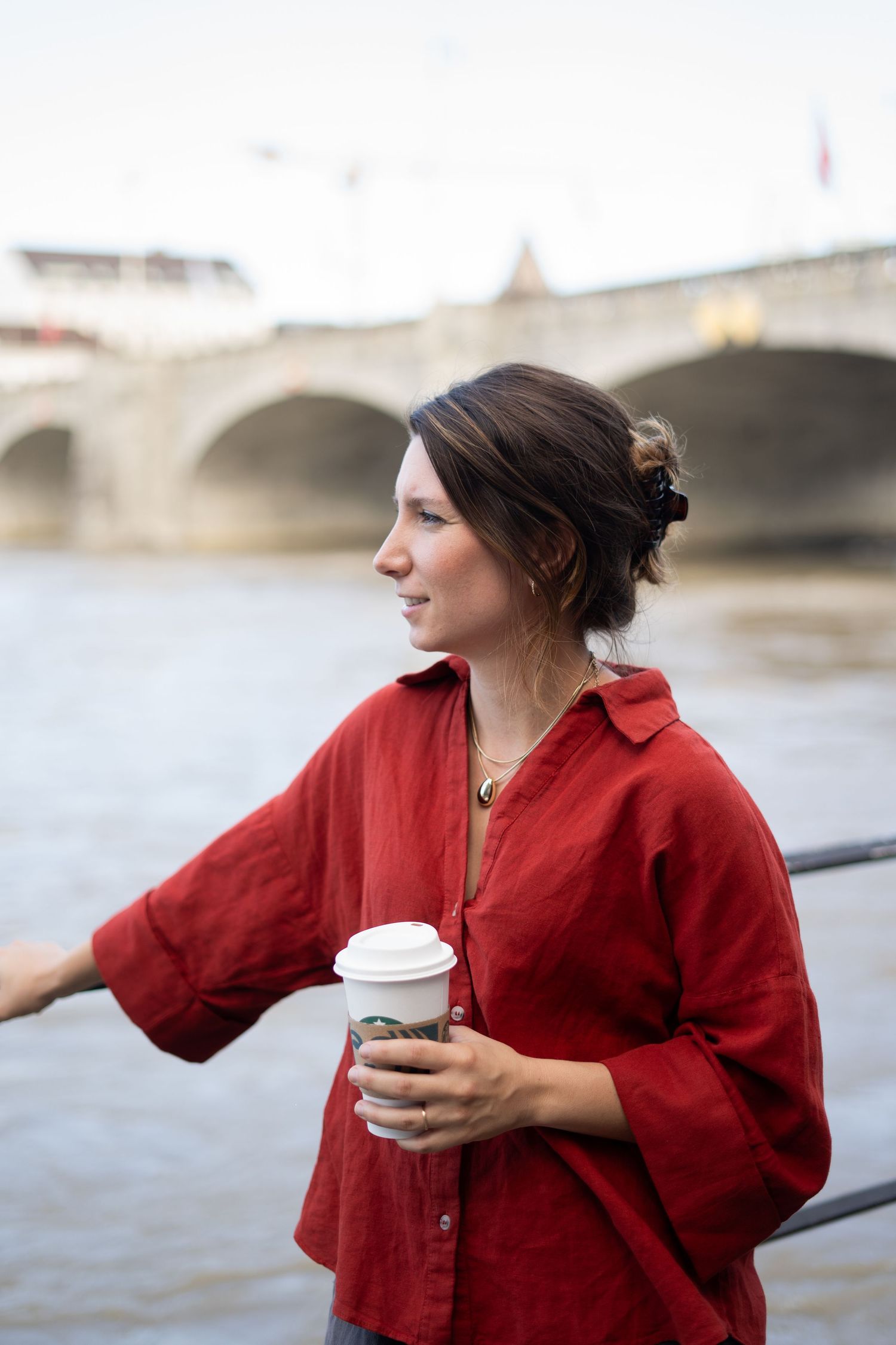 Person in red shirt holding coffee cup while standing by river with bridge in background.