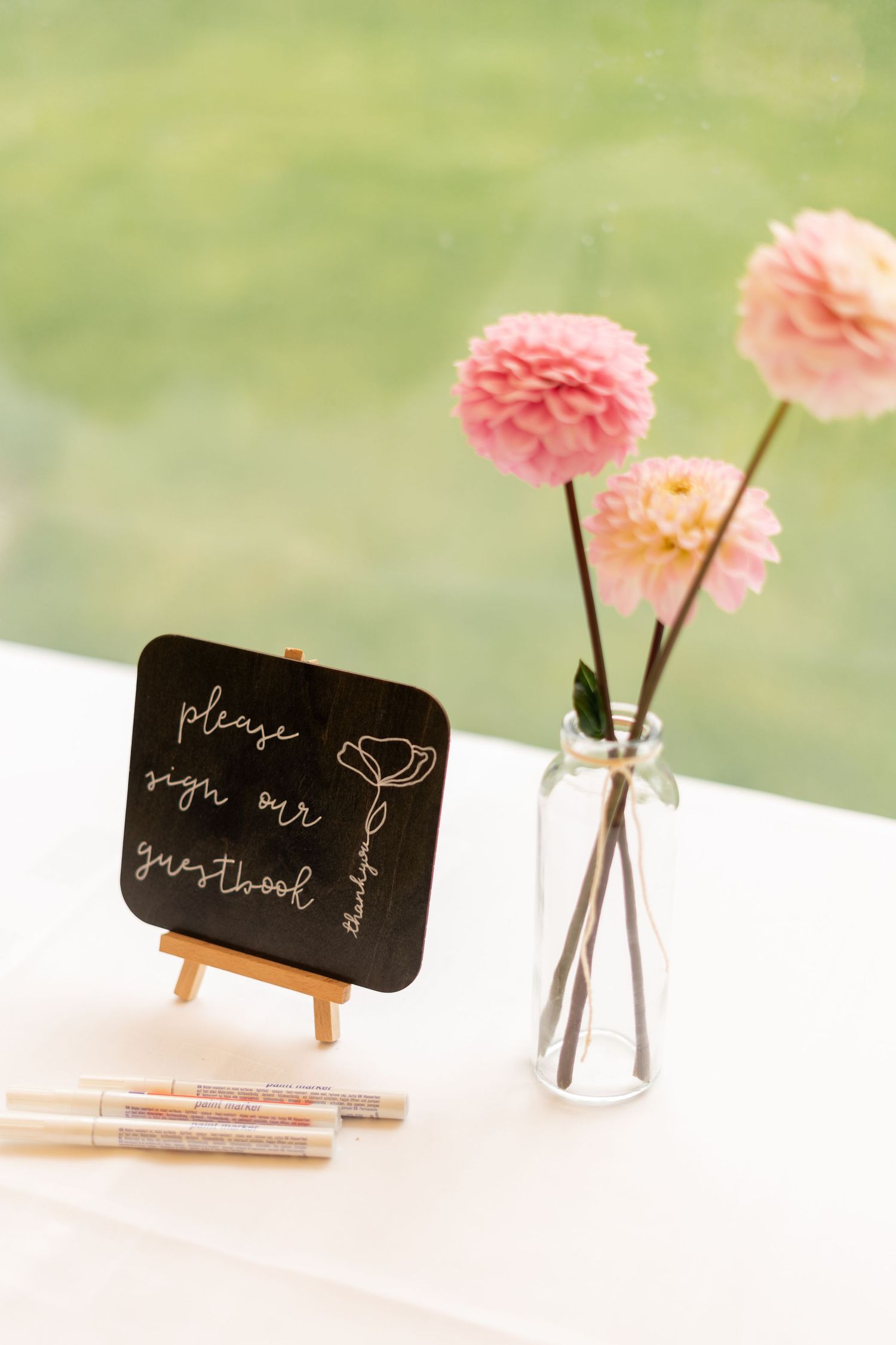 Pink carnations in a clear vase next to a chalkboard sign on white table with blurred green background.