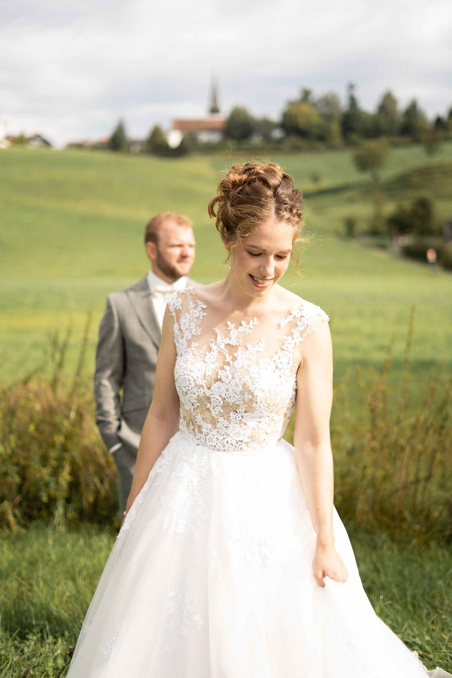 A wedding couple poses in a scenic green field with a church visible on a distant hilltop.
