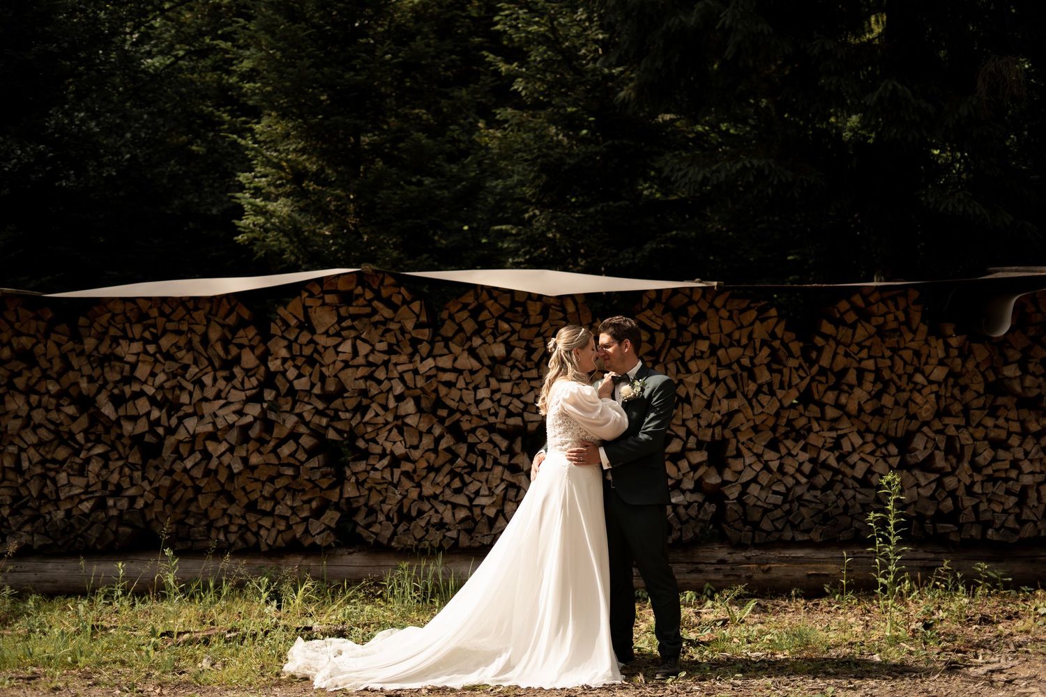 A romantic wedding couple embraces in front of stacked firewood during a rustic outdoor photoshoot.