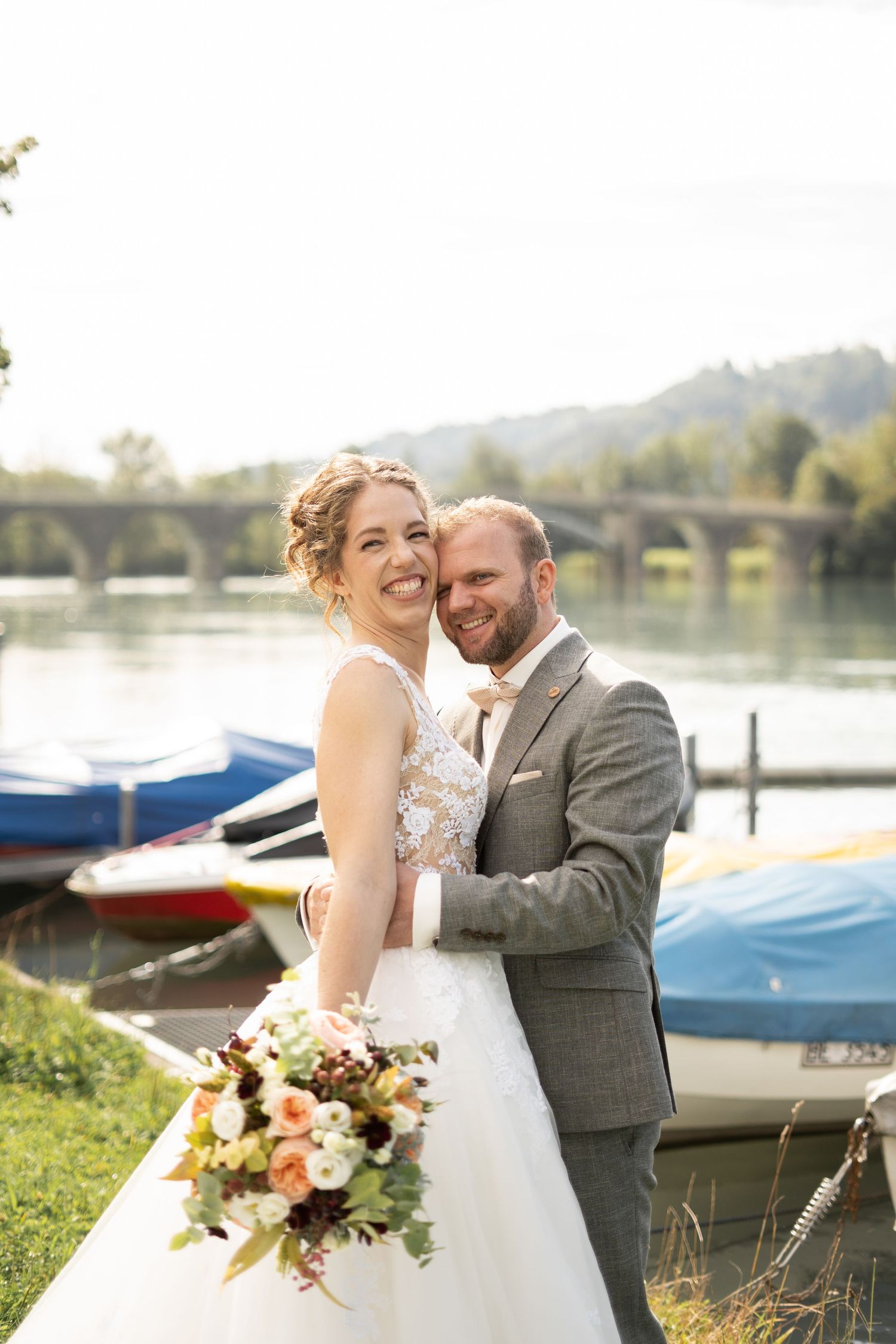 A wedding couple poses by colorful boats on a lake with an arched bridge in the background.