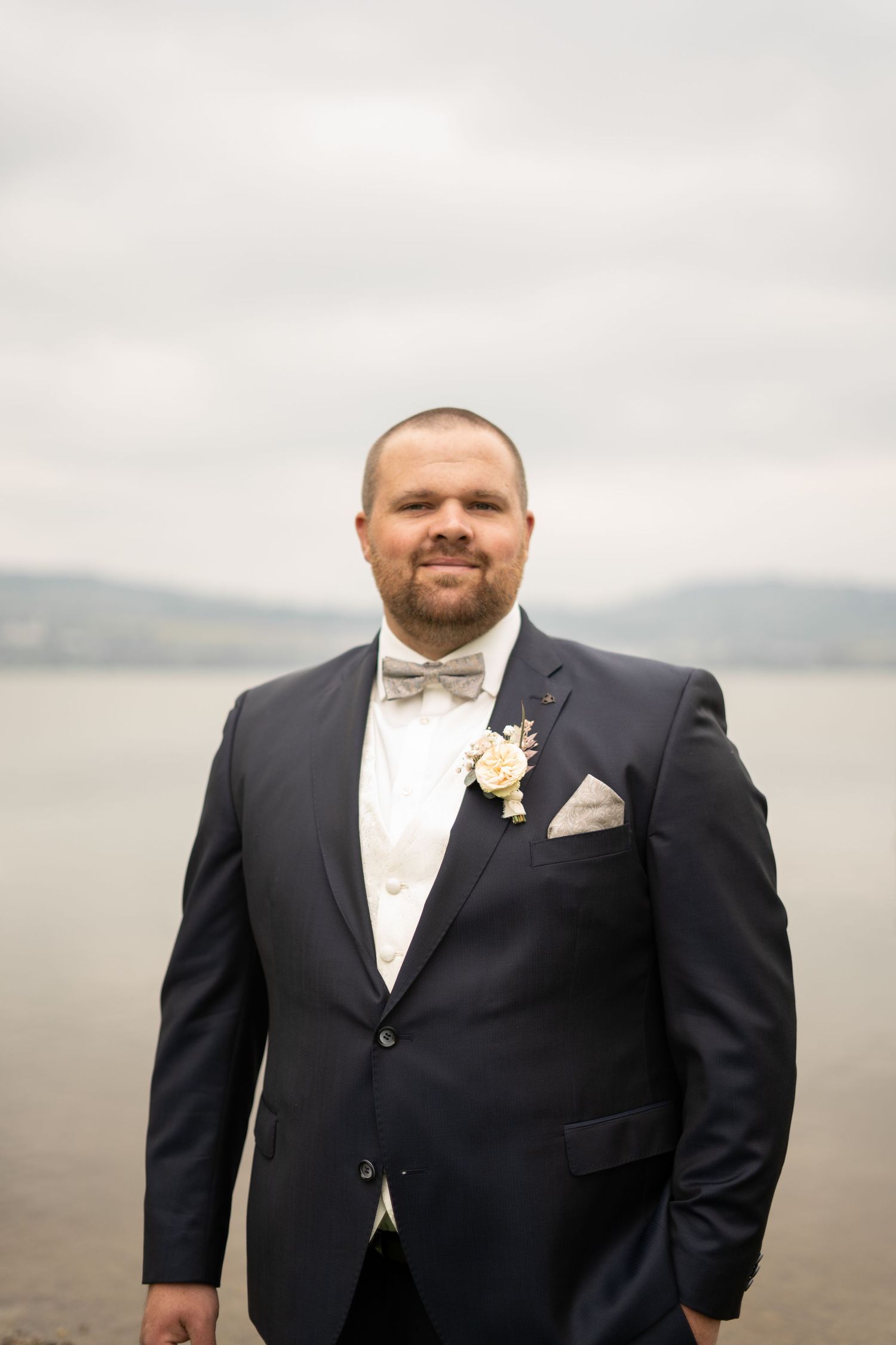 A wedding photo of a groom in a classic black tuxedo and bow tie with a floral boutonniere against an overcast sky.