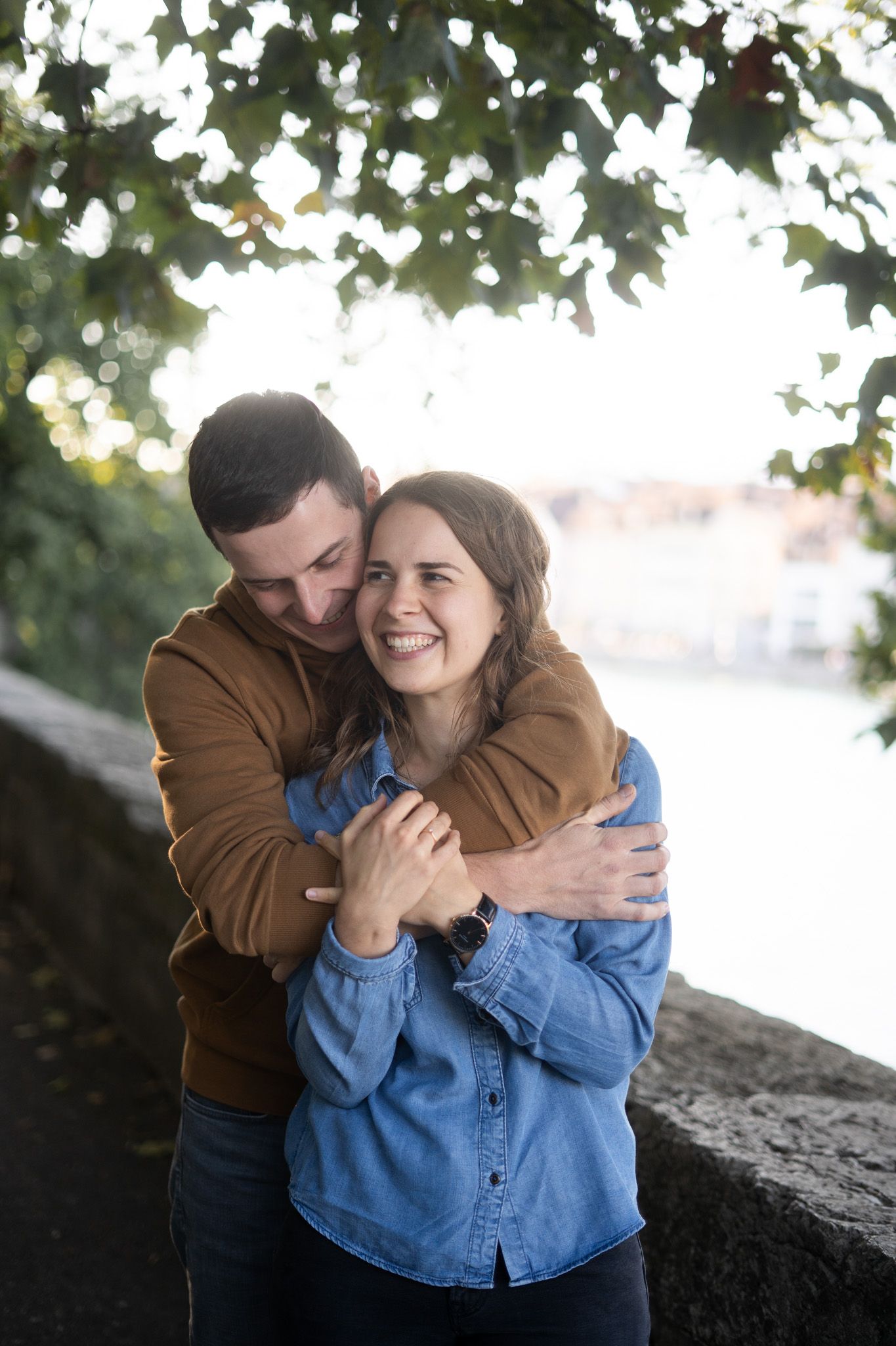 A romantic moment under tree leaves as couple embraces and shares laughter by the water.