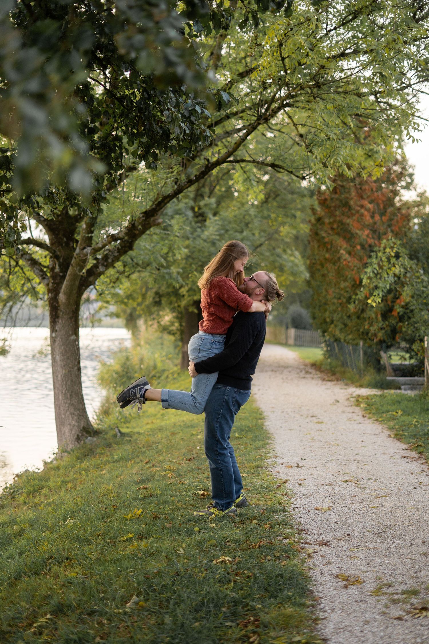 A joyful couple shares a romantic lift moment on a tree-lined path near water.