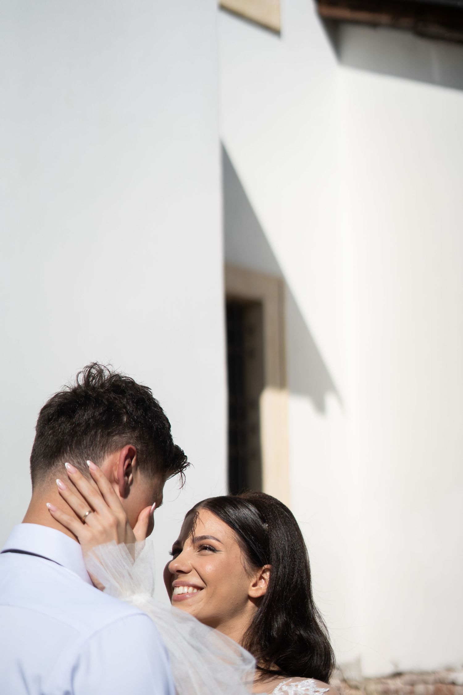 Joyful wedding couple shares intimate moments against a white wall in natural sunlight.