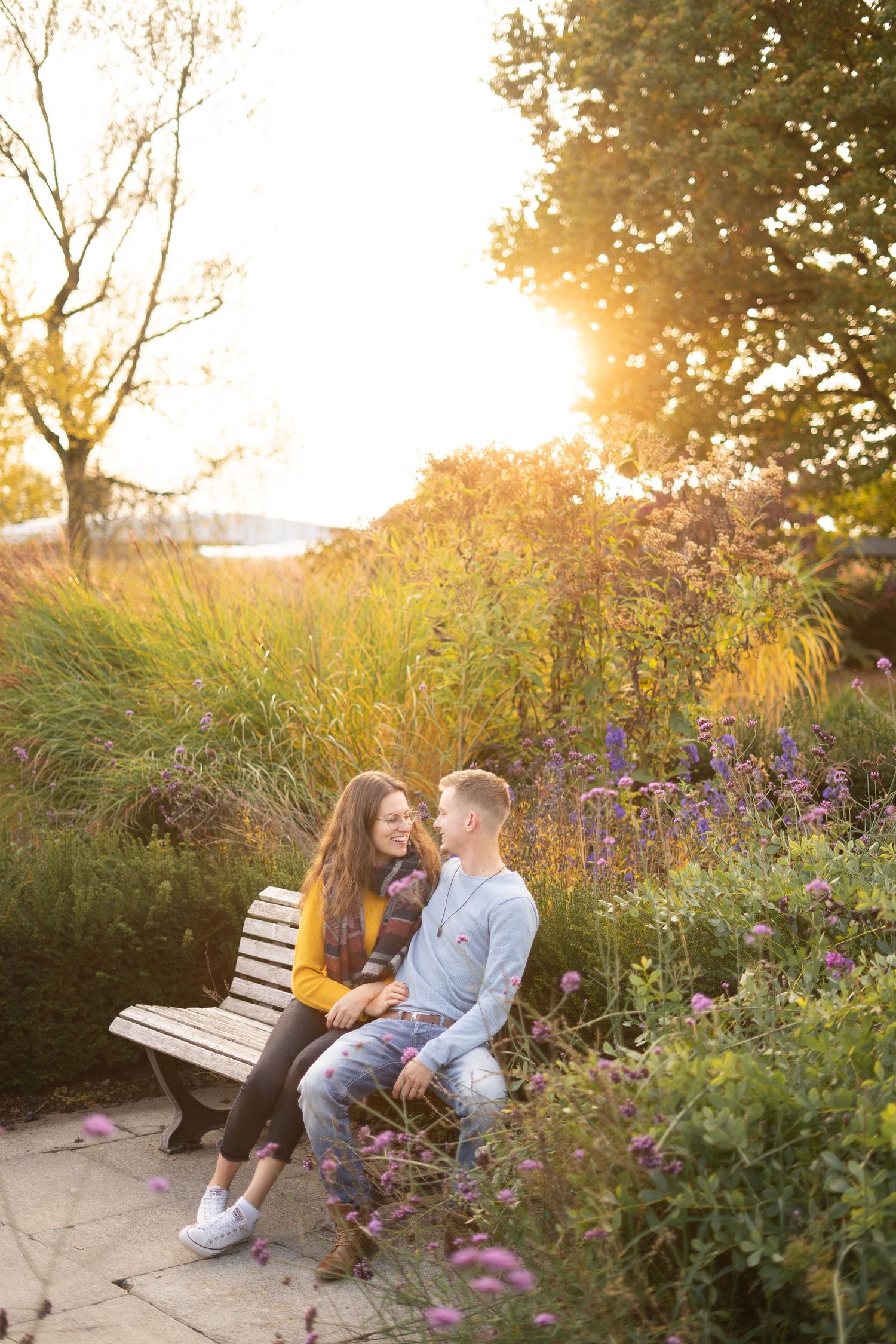 A couple shares a tender moment on a park bench surrounded by purple wildflowers at sunset.