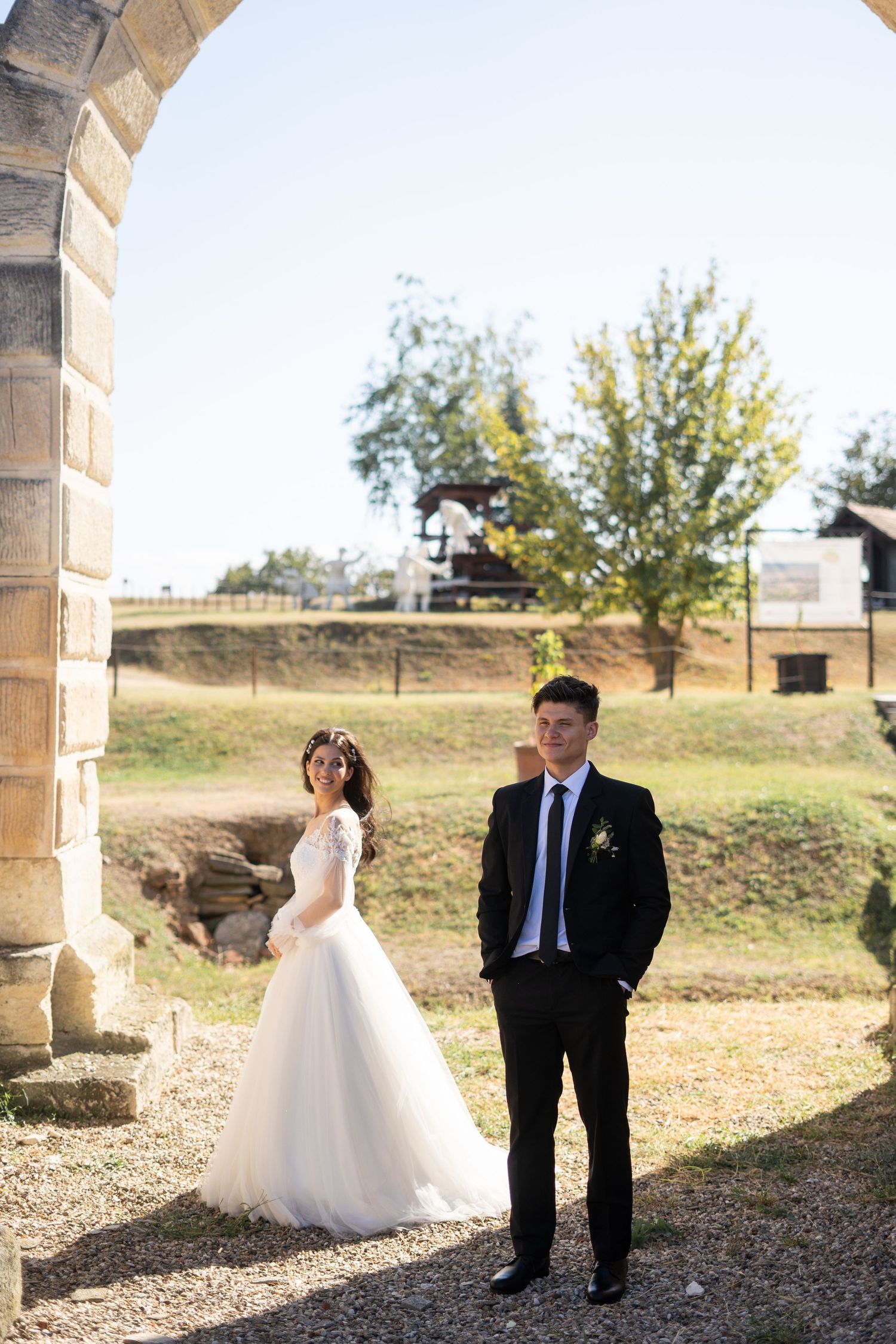 A wedding couple sharing intimate moments under an outdoor stone colonnade on a sunny day.