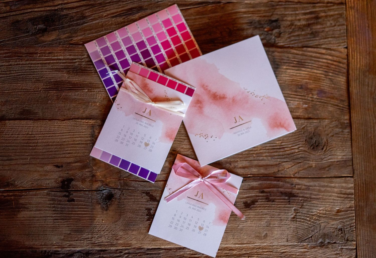 Wedding stationery with pink watercolor design and ribbon details on wooden surface.