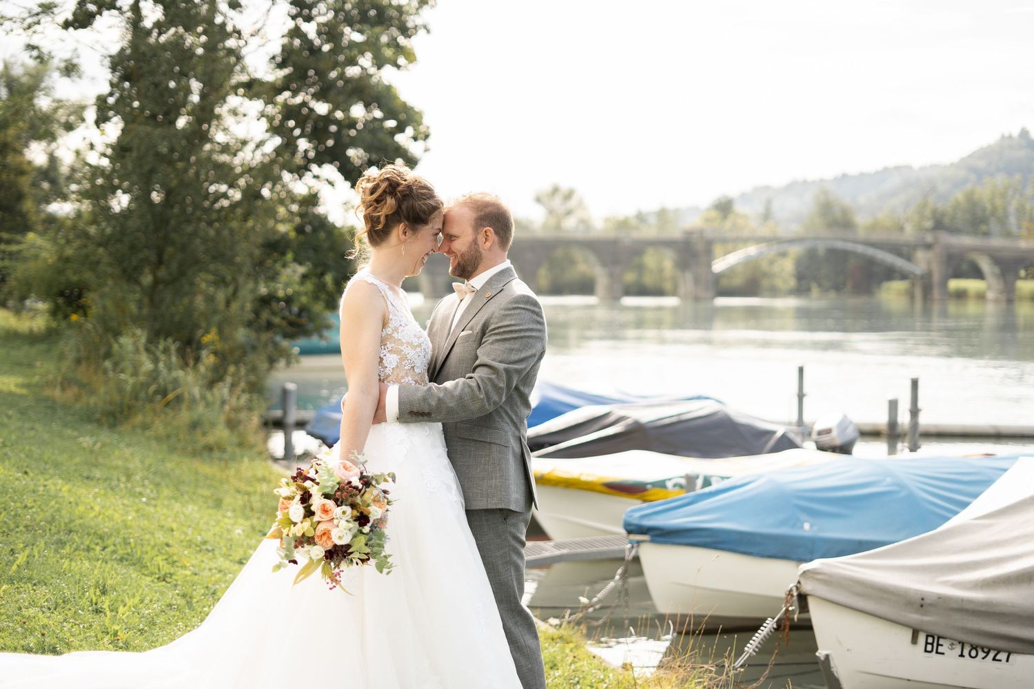 Newlyweds share a tender moment near docked boats along a lakeside.