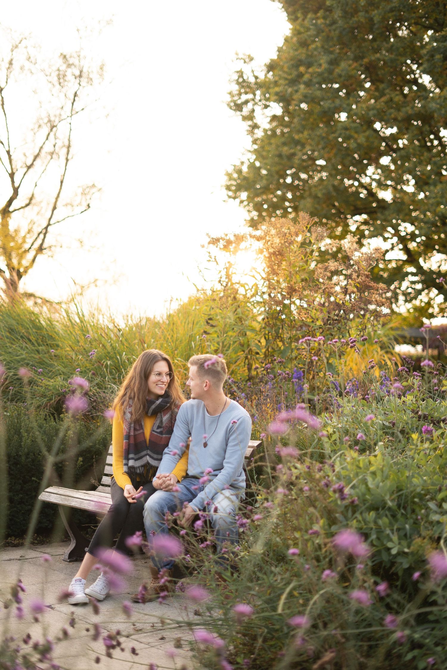 Two people sharing romantic moment on bench amid purple flowers at sunset.