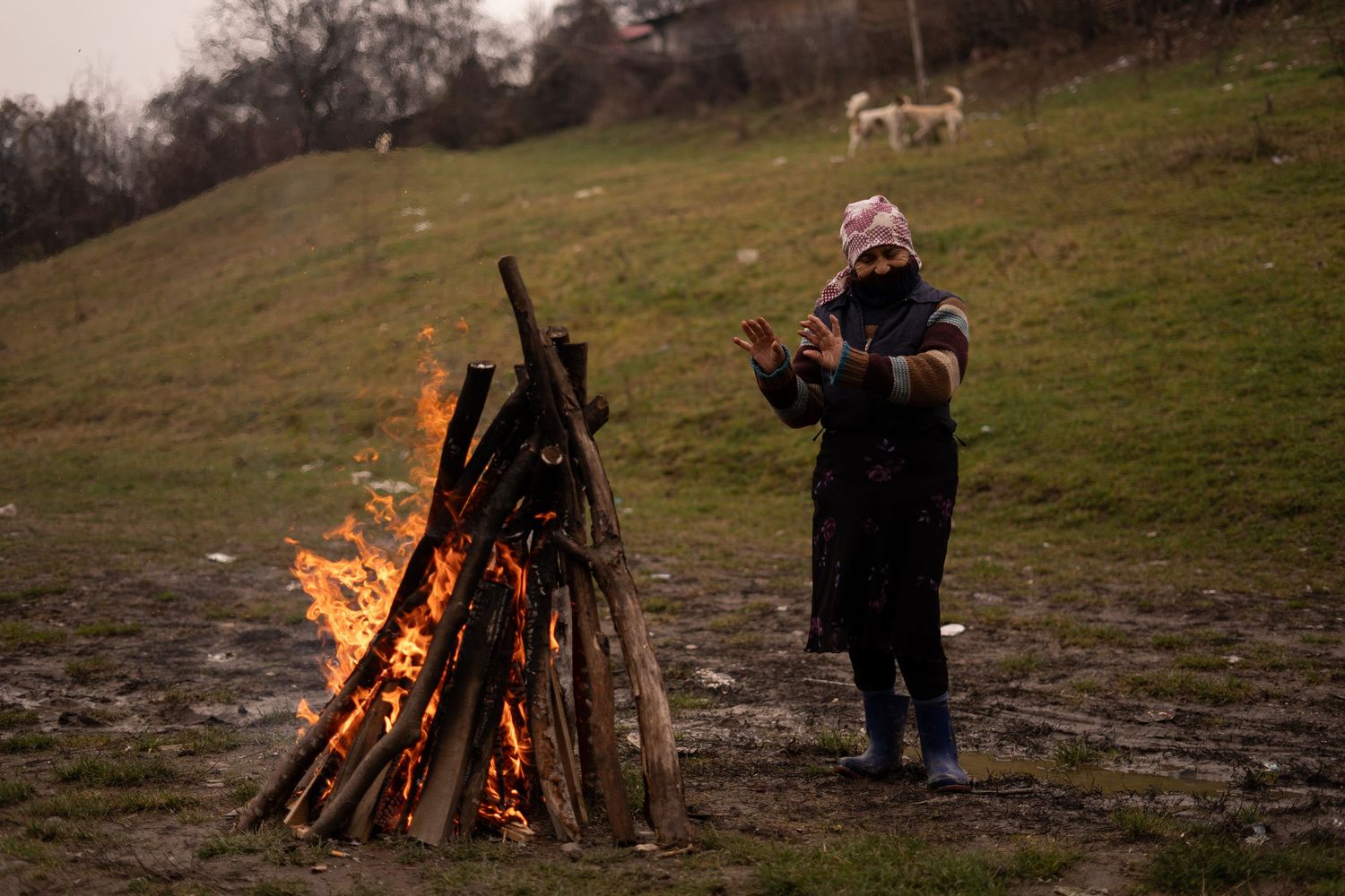 A warmly dressed figure stands beside a large bonfire on a hillside at dusk.