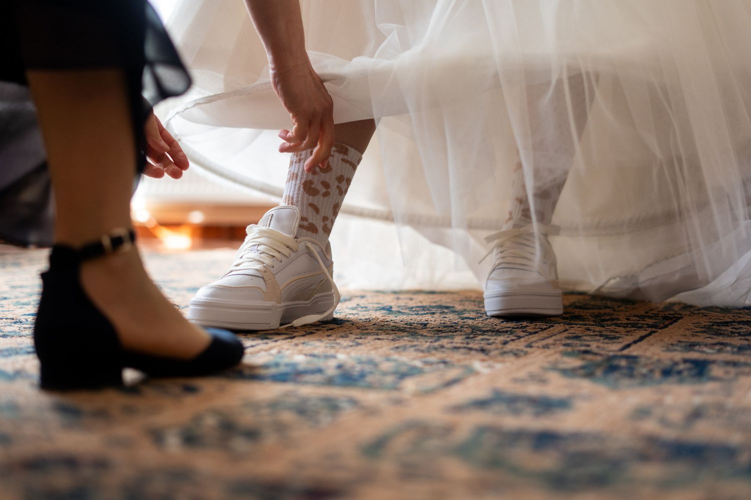 Close-up of white sneakers being put on under a wedding dress on a patterned carpet.