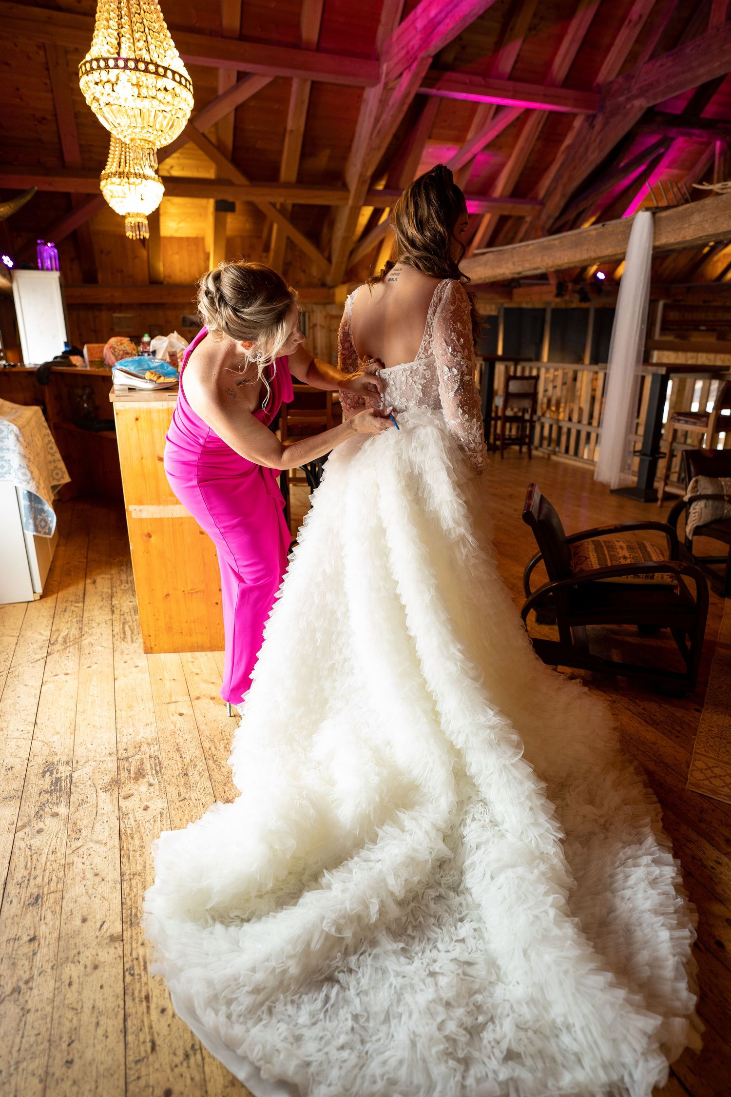 Wedding dress being adjusted in rustic barn venue with chandelier and pink lighting.