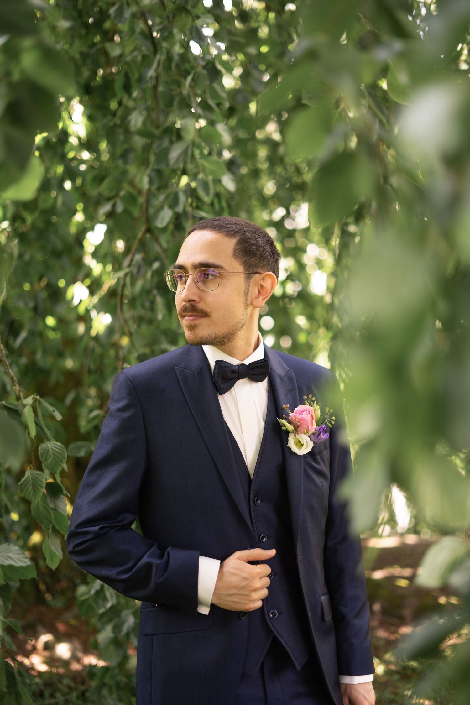 Groom in navy suit with bowtie stands under leafy green branches in natural lighting.