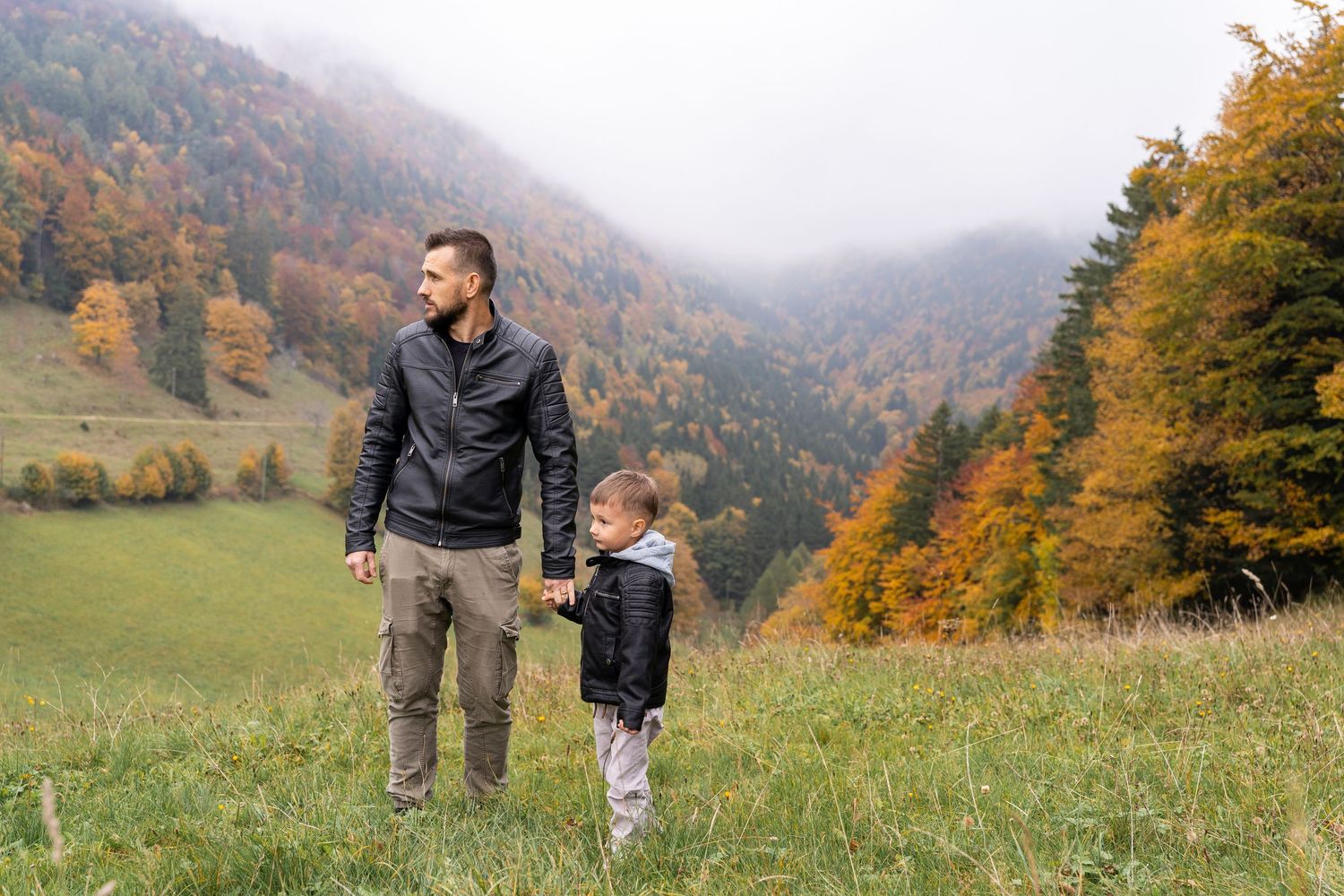 Two people stand in a grassy meadow surrounded by autumn mountains with colorful fall foliage in the background.
