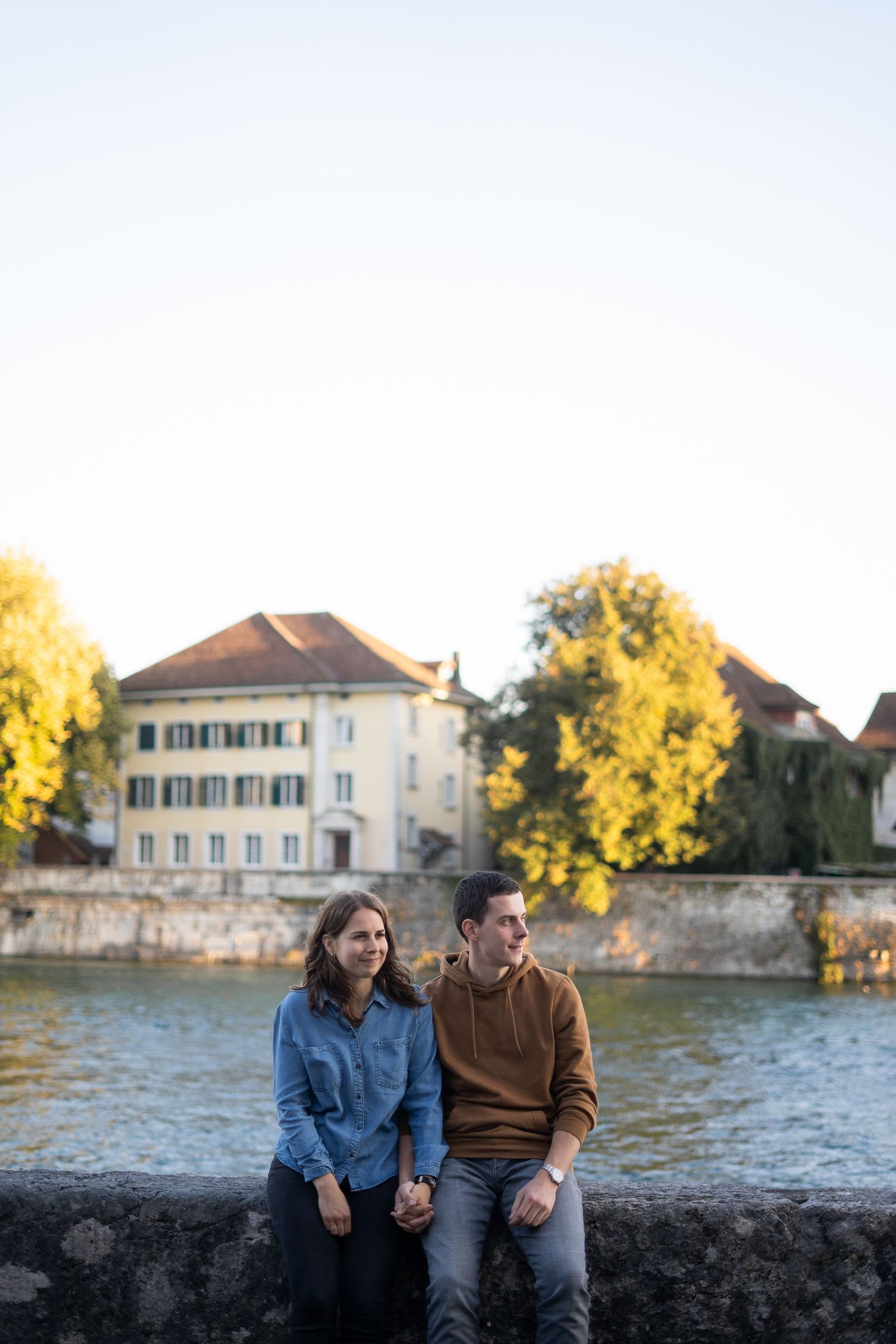 Couple sitting together on stone wall with historic building and autumn trees in background.