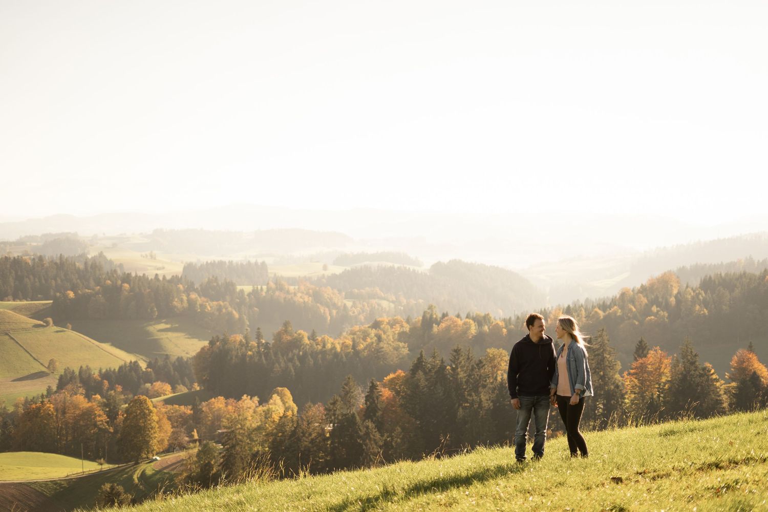 A couple stands together on a grassy hilltop overlooking a scenic autumn valley.