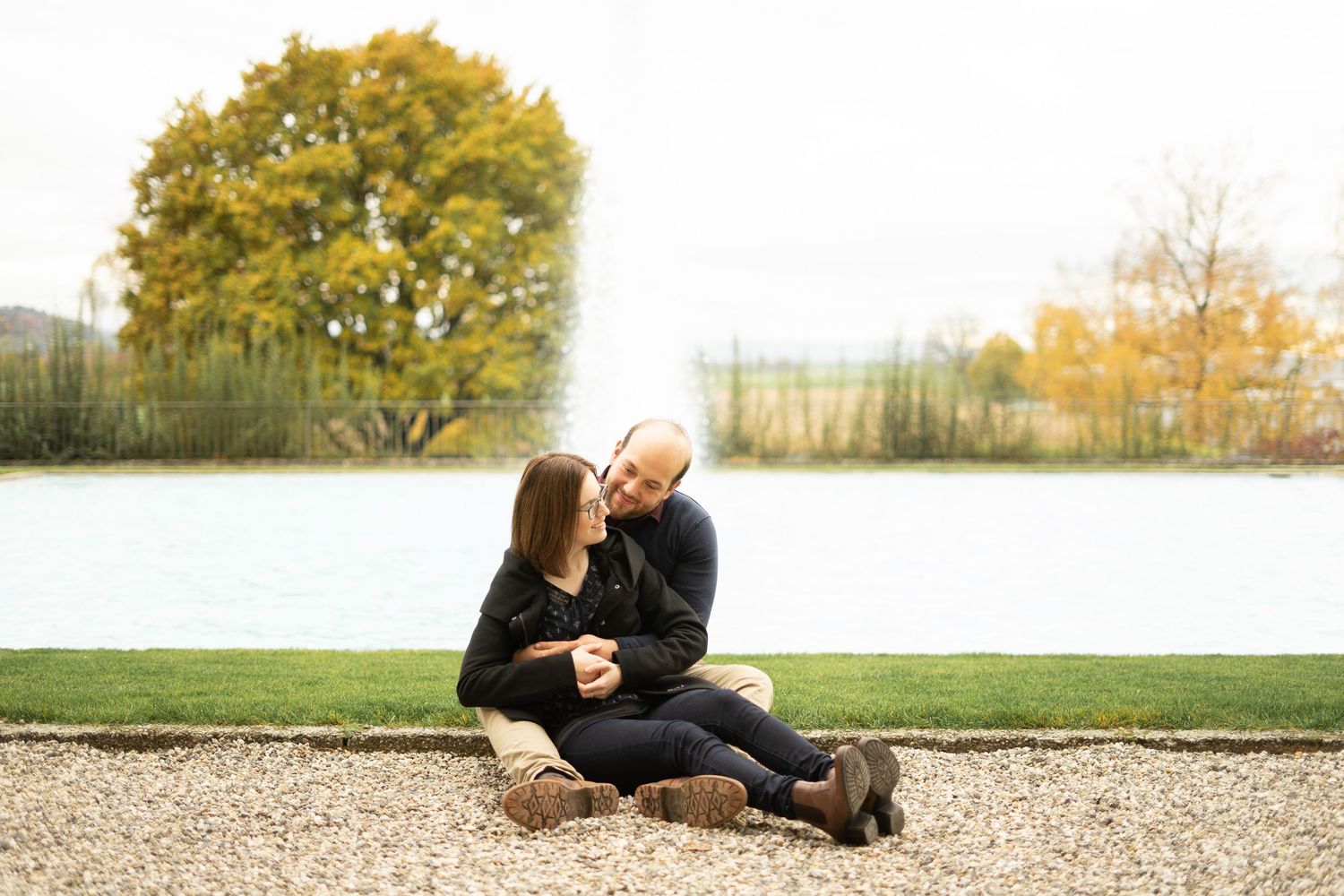 A couple sits together on gravel path near a lake during fall season.