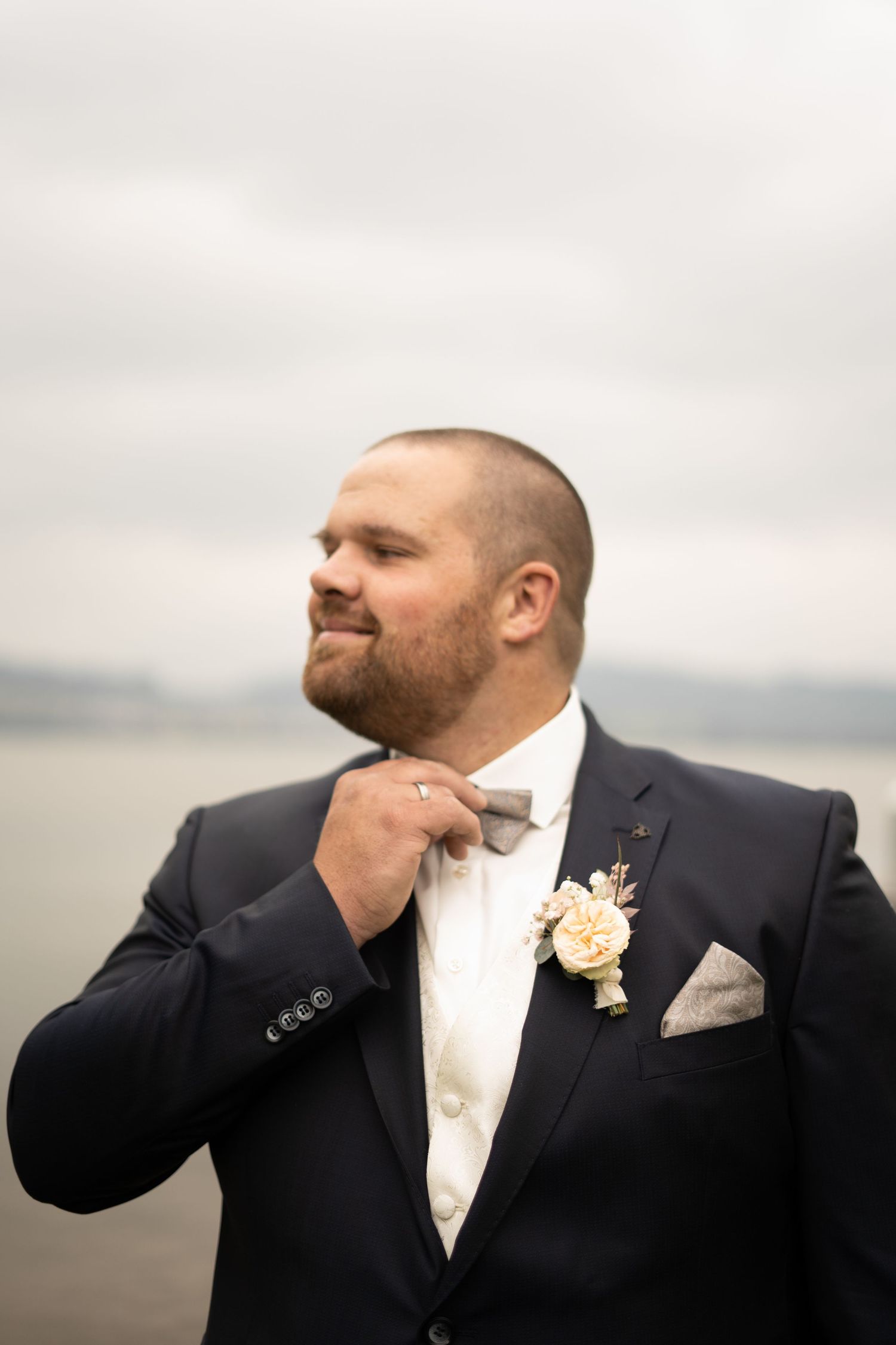 A groom adjusts his bow tie while wearing a black tuxedo with a boutonniere against a cloudy background.