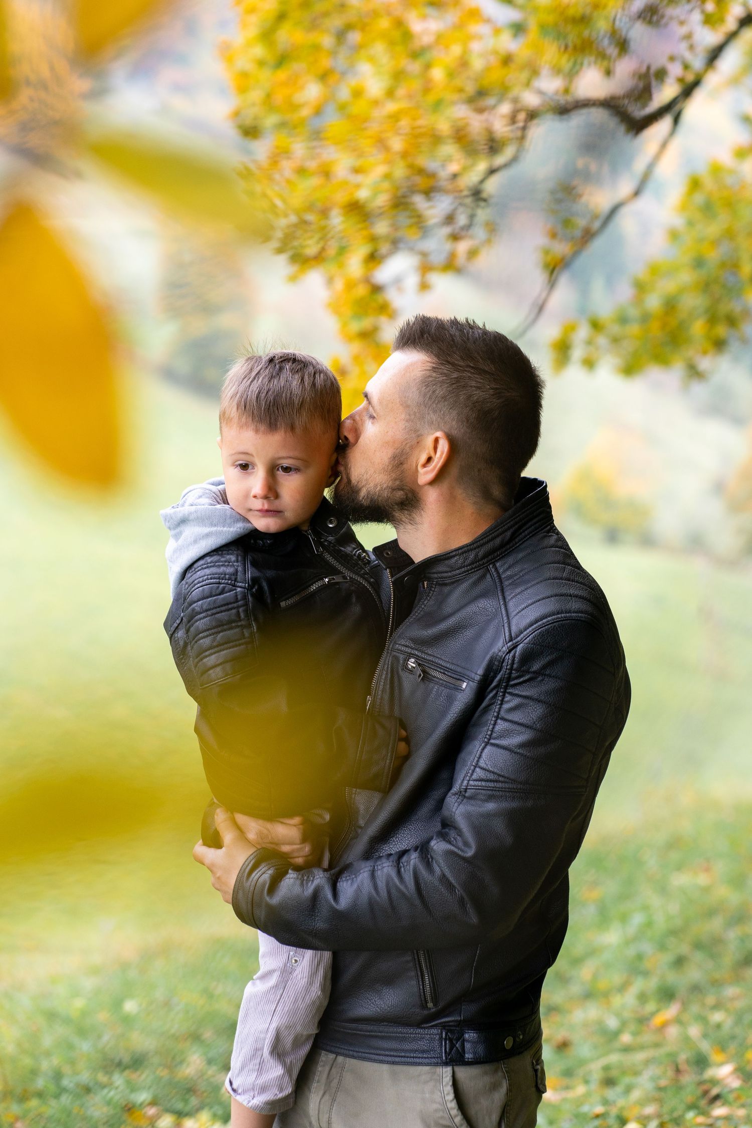 A tender autumn moment captured between parent and child in a park with golden yellow leaves in the background.