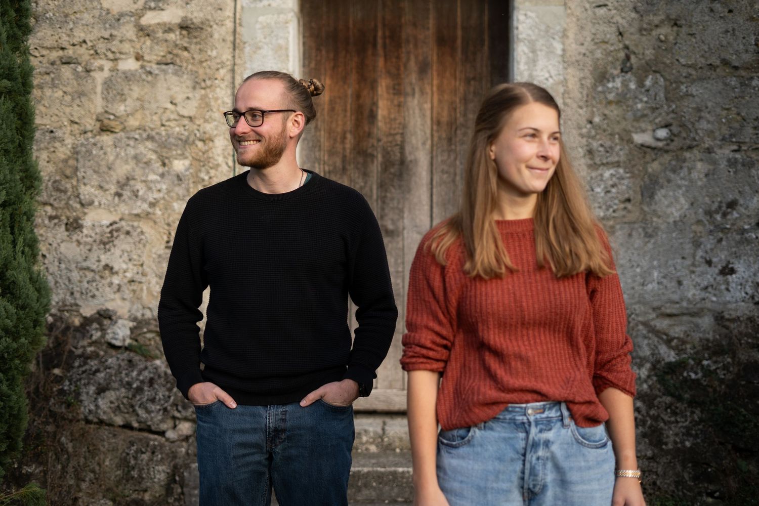 Couple sitting by rustic stone wall in casual outfits during outdoor photoshoot.