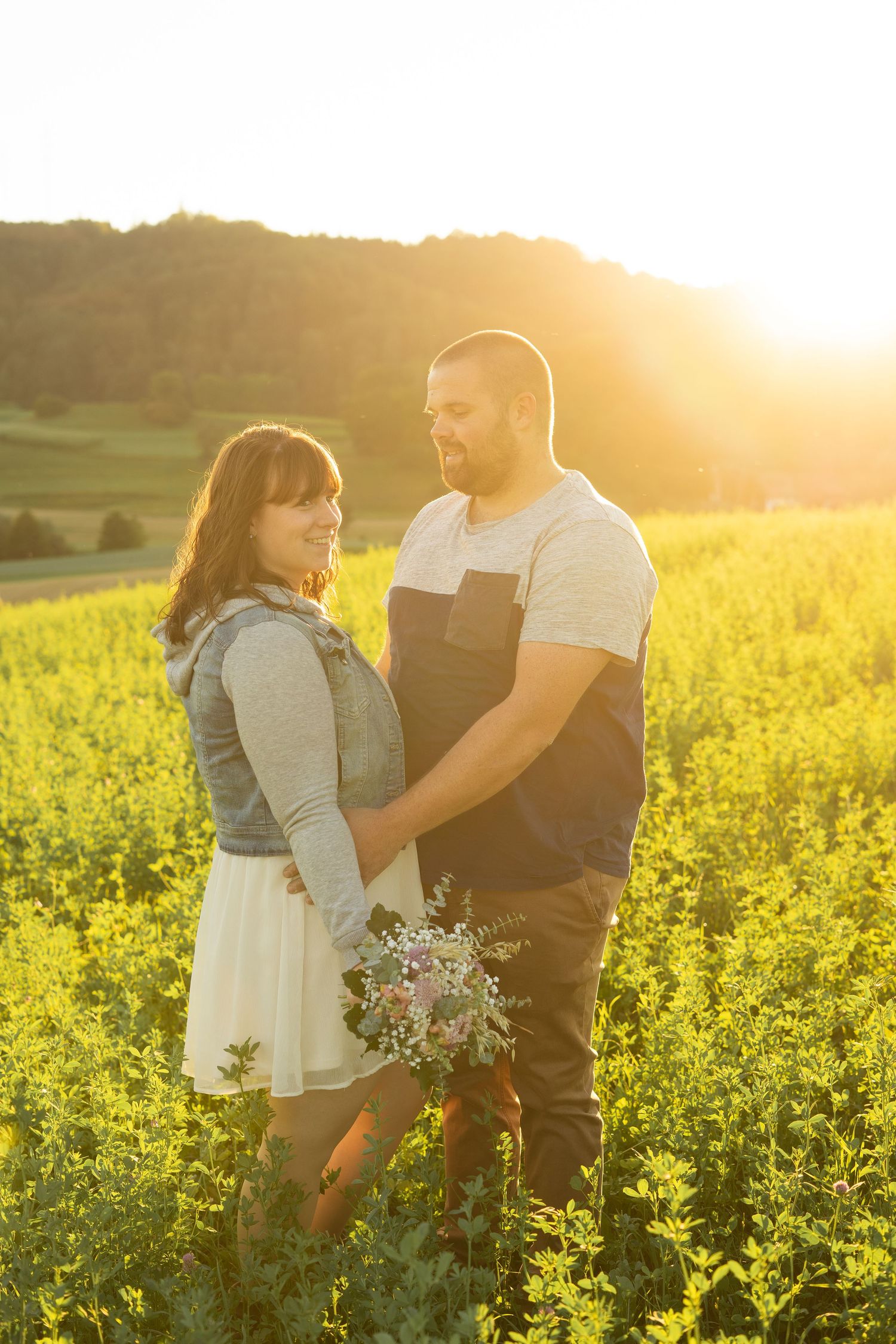 A couple poses in a sunlit yellow canola field at golden hour holding a wildflower bouquet.