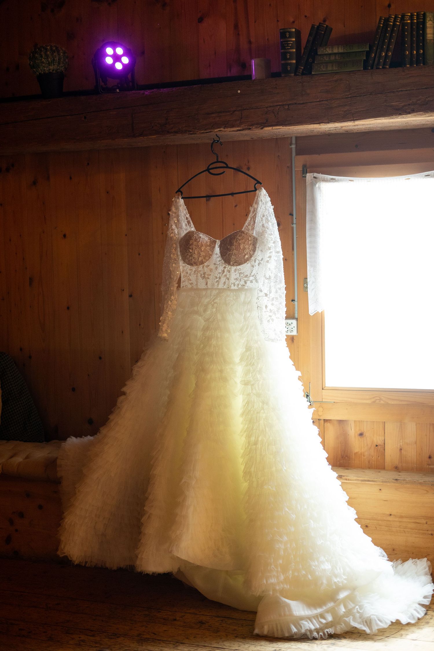 Elegant white lace wedding dress hanging in rustic wooden room with natural window light.