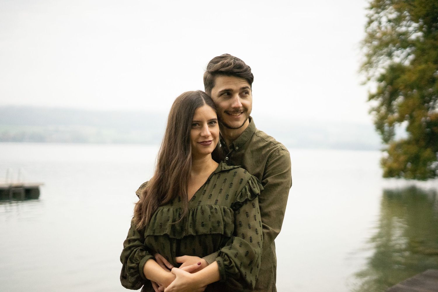 A couple in matching olive-colored outfits embrace by a misty lake.