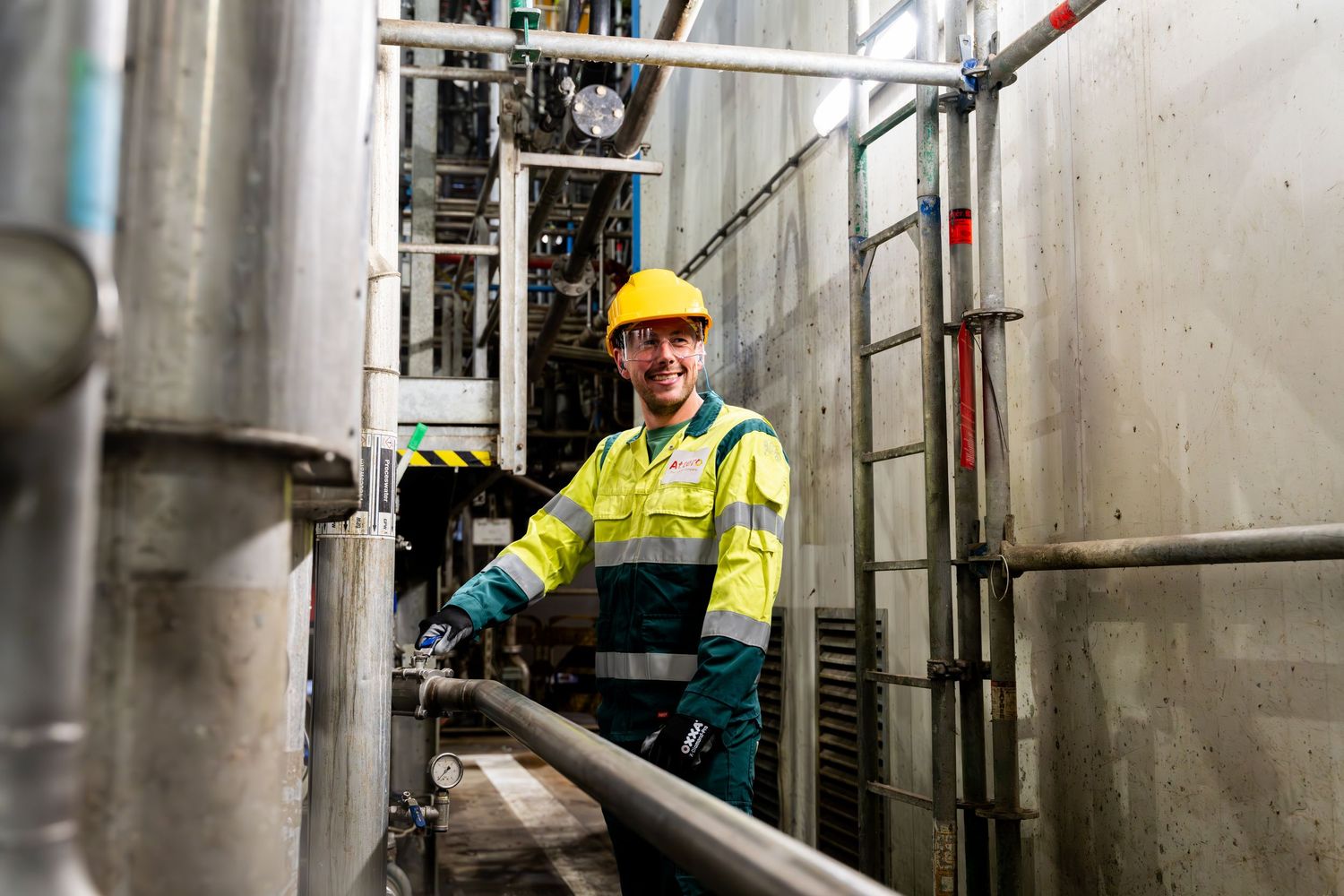 Industrial worker in safety gear inspects equipment in a manufacturing facility with metal tanks and pipes. - Noelle Schmeltz Fotografie