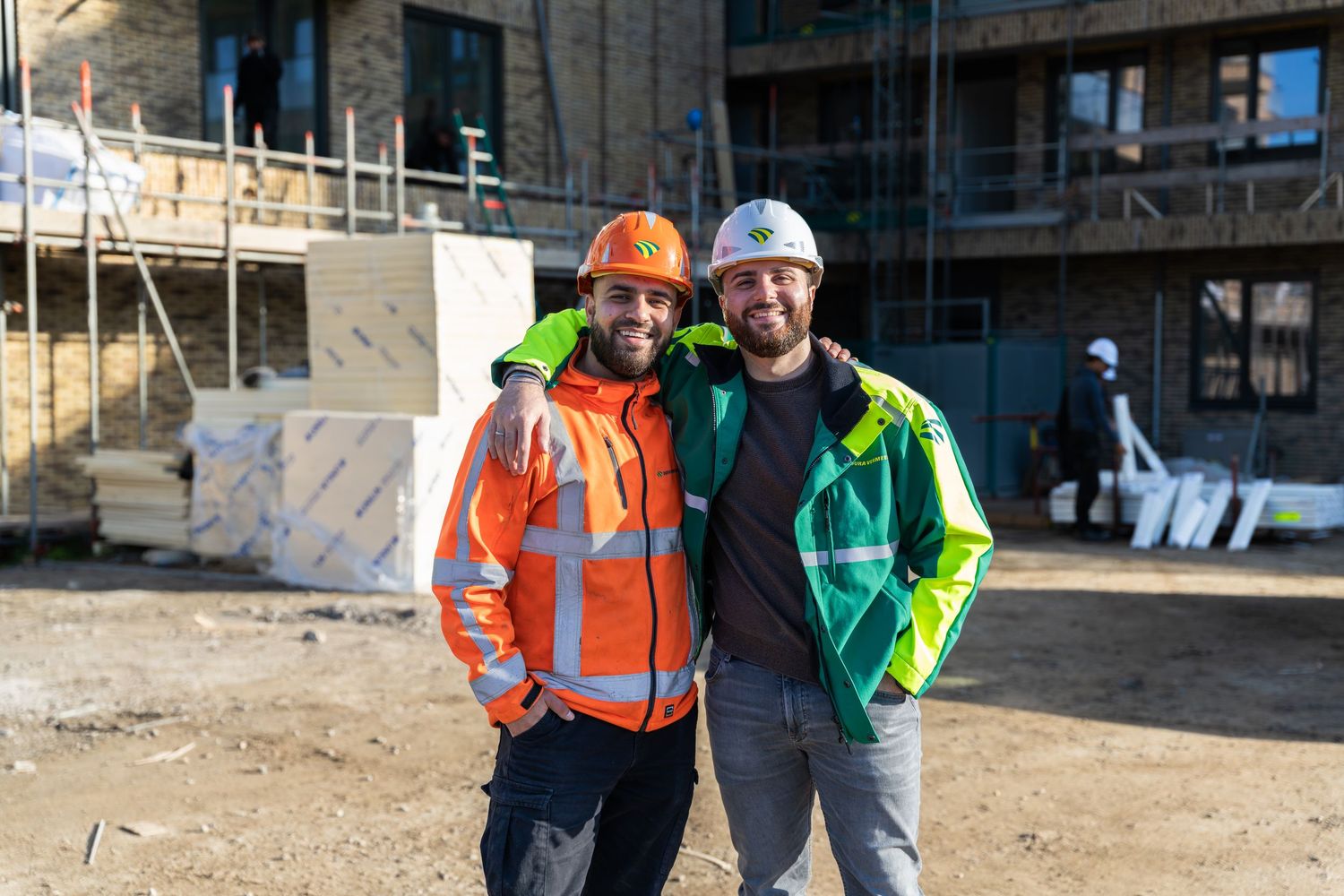 Two construction workers in safety gear and hard hats stand together at a building site.